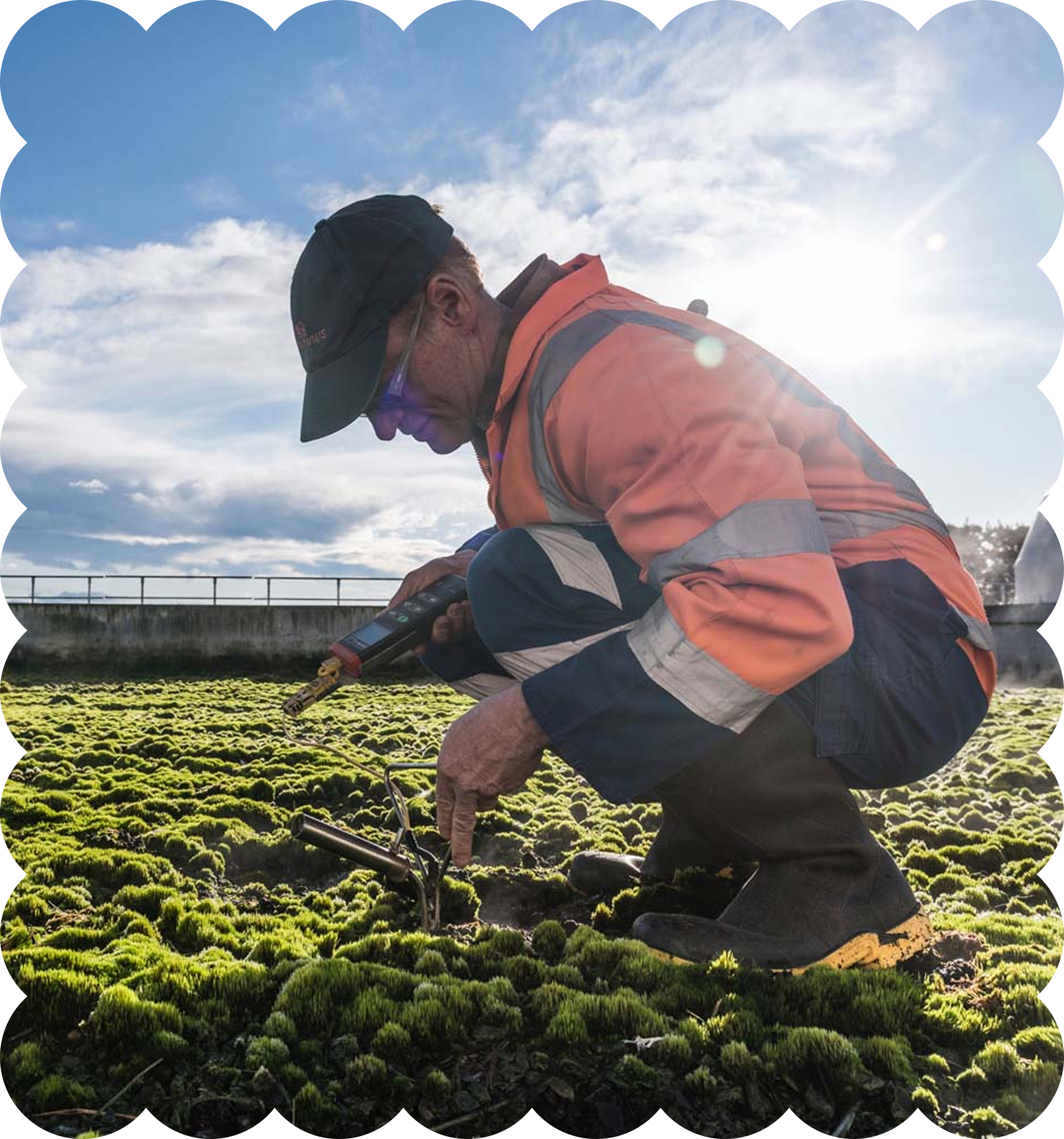 A man in an orange safety jacket and black cap kneeling and working with gardening tools in a green moss landscape, with a bright blue sky and white clouds in the background.