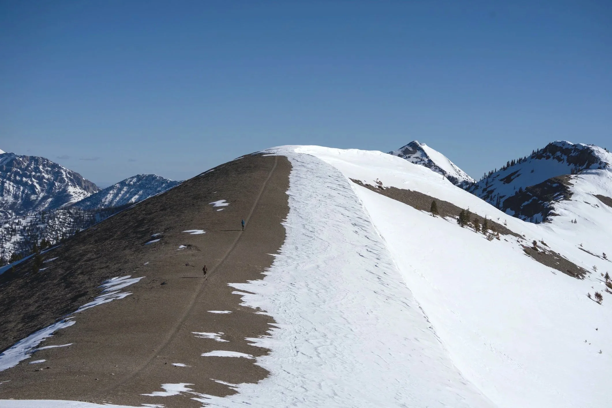 Snow-capped mountain peaks under a clear blue sky with a snow-covered ridge and a trail with a few hikers walking on it.