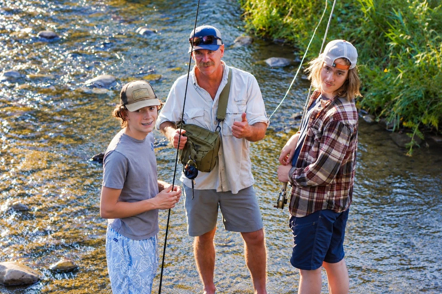 Three people standing in a shallow, rocky river with fishing rods, surrounded by green vegetation on the riverbank during daytime.