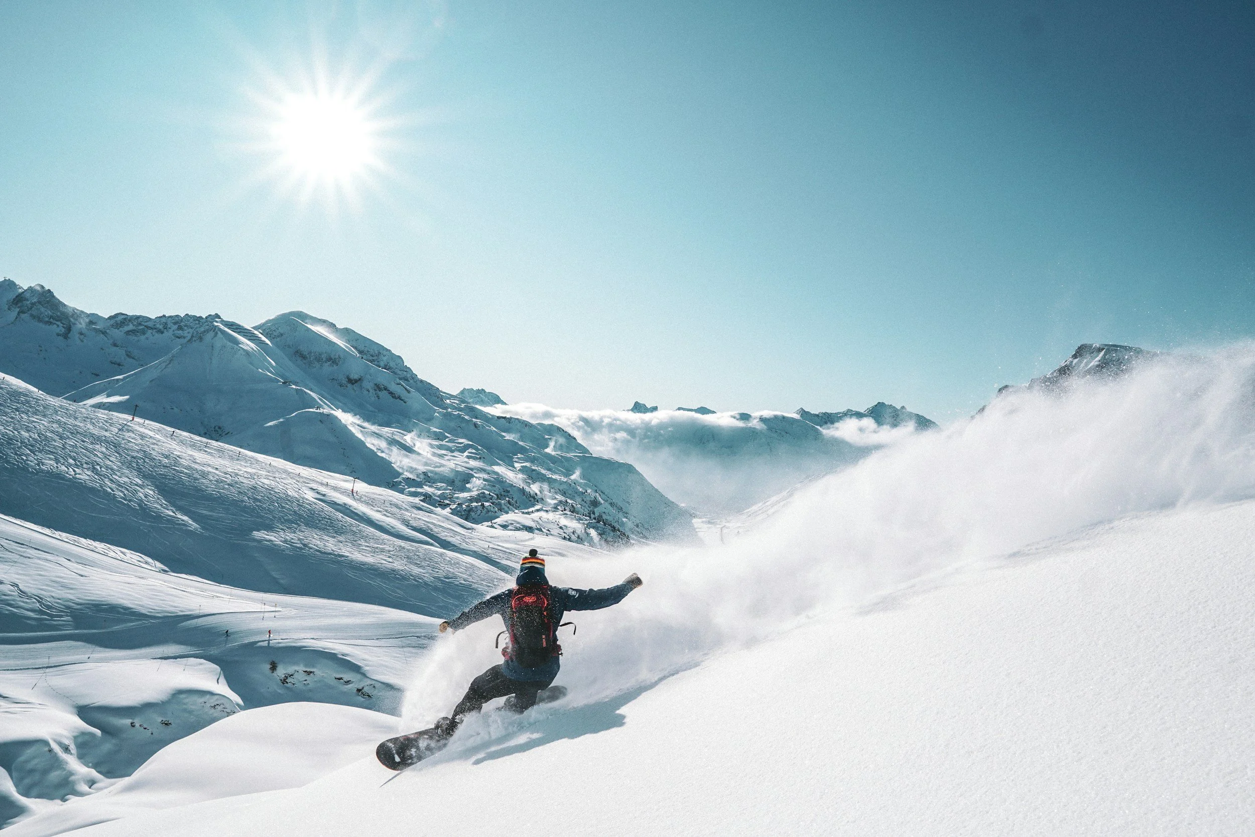 A person snowboarding down a snowy mountain slope under a bright sun, with snow-covered peaks in the background and a clear blue sky.