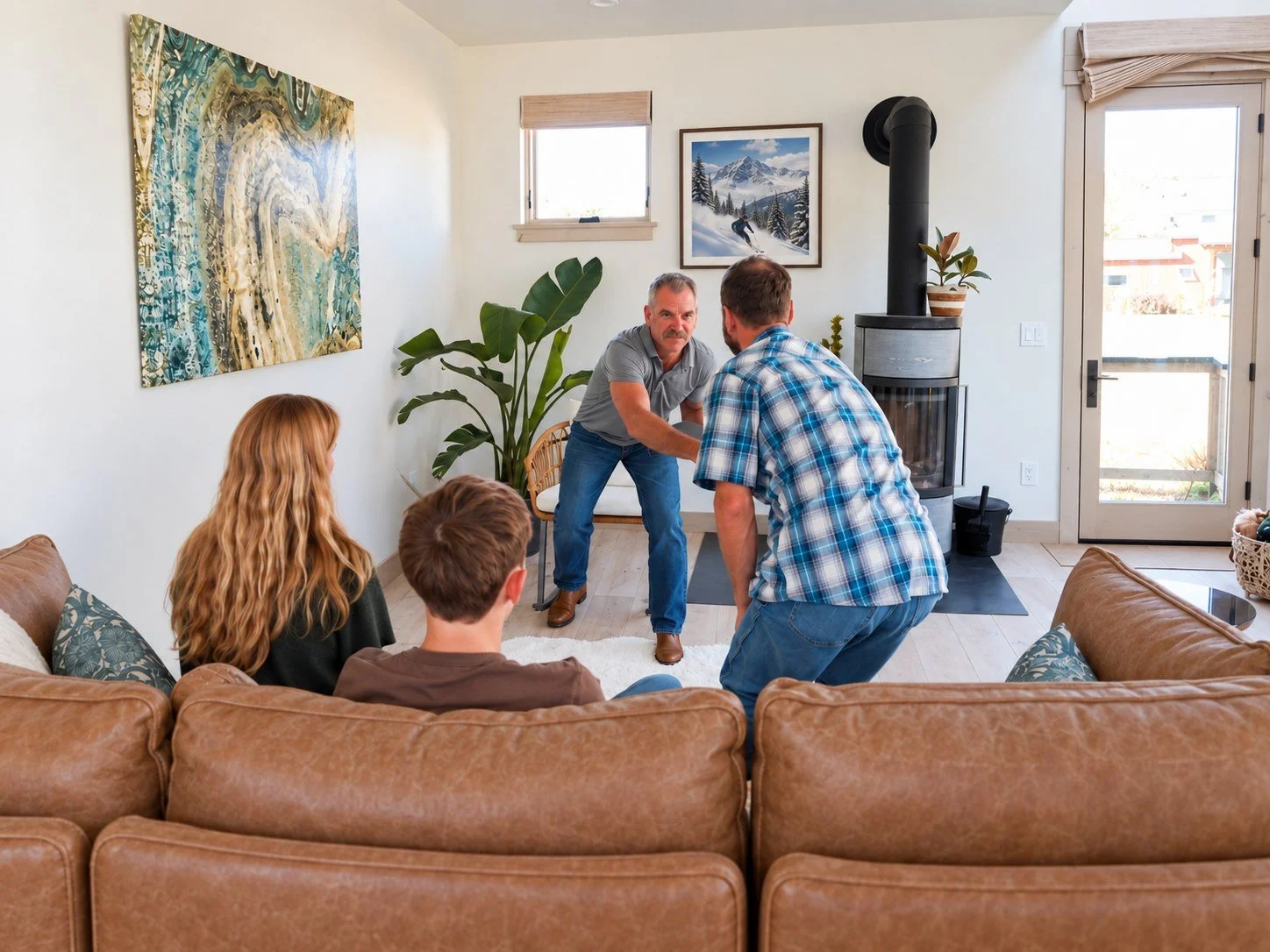 Four people sitting in a living room, watching a man in a gray shirt demonstrate a self-defense move. The living room features artwork, plants, a wood stove, and a glass door.