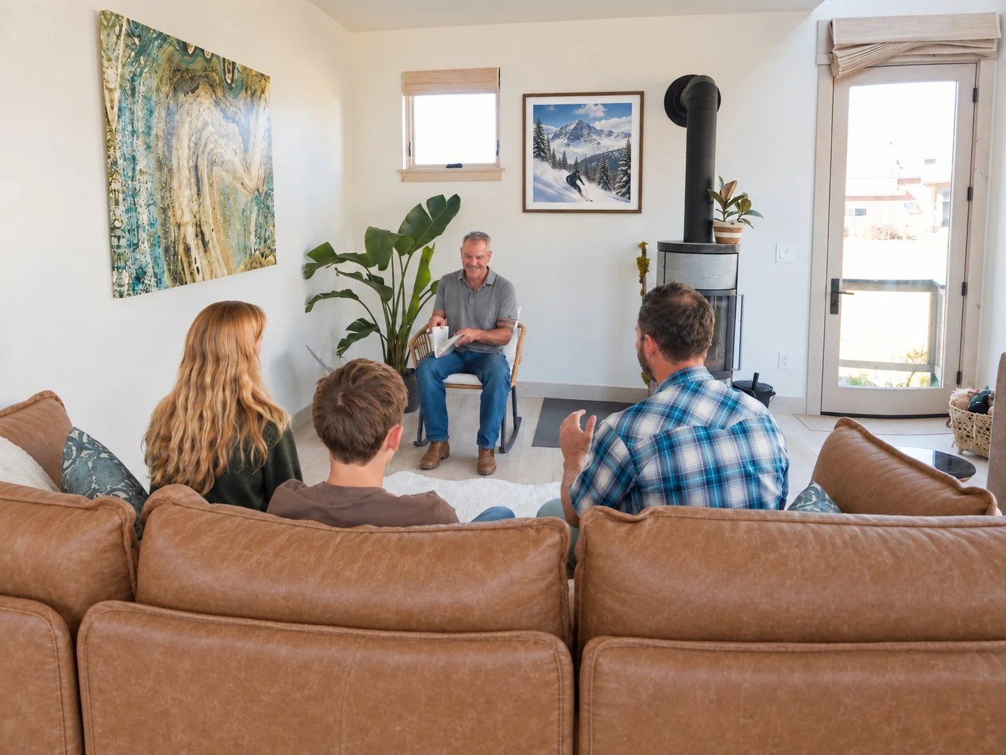 A man sitting in a chair leading a group therapy session or support group in a bright, cozy living room. Four people are seated on a tan leather couch listening attentively. The room has a large window, art on the walls, a potted plant, and a wood stove.
