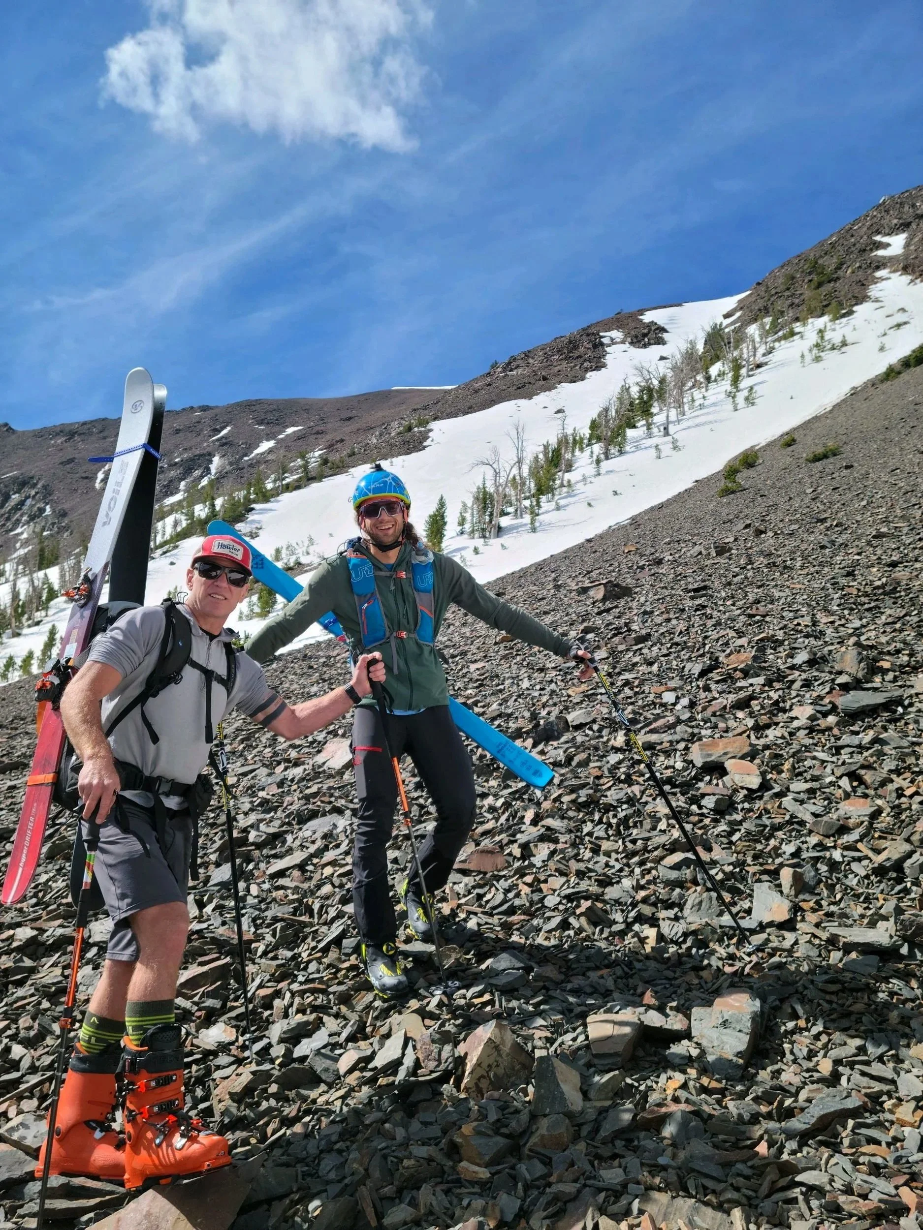 Two men in outdoor gear, including helmets, sunglasses, and backpacks, stand on rocky mountain terrain with snow patches and sparse trees, holding ski poles, with a partly cloudy sky overhead.