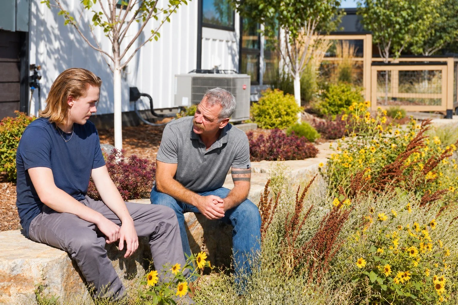 A young man and an older man sit on a stone ledge in a garden, having a conversation. The garden has yellow and purple flowering plants, small trees, and some residential and outdoor structures in the background.