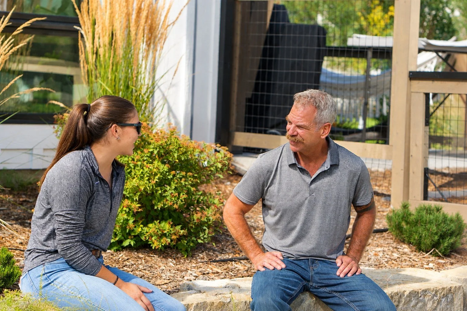 A young woman and an older man are sitting outdoors on a stone wall, engaged in conversation, with greenery and a fence in the background.