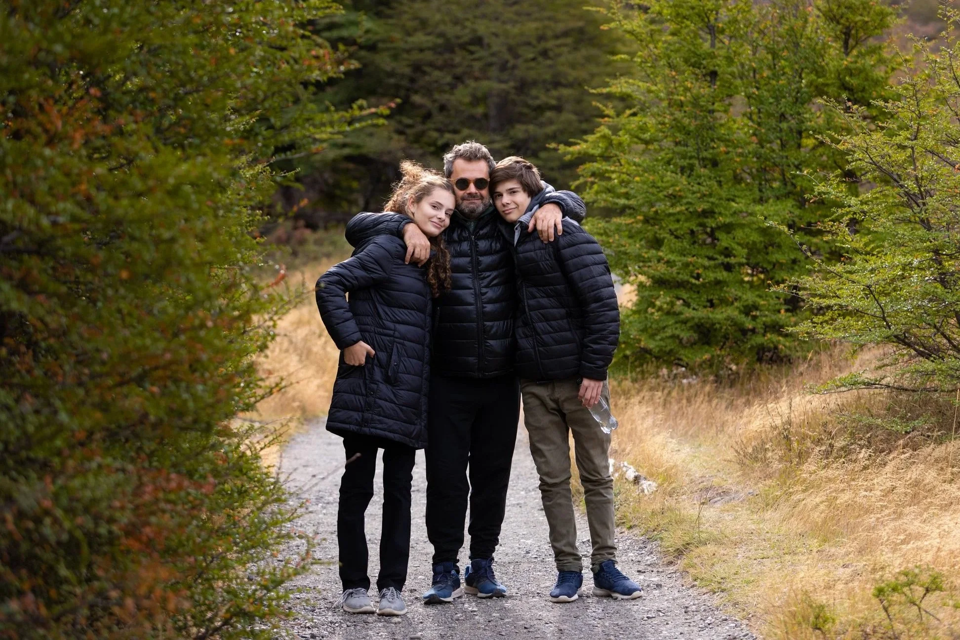 A family of three, two children and an adult man, standing on a dirt path outdoors surrounded by green trees and foliage, with the adult hugging both children.
