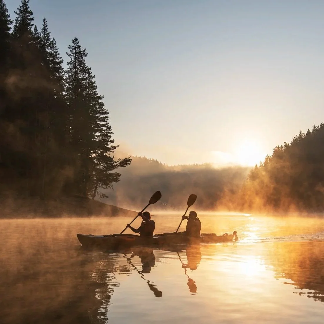 Two people kayaking on a misty river at sunrise or sunset, surrounded by forested hills.