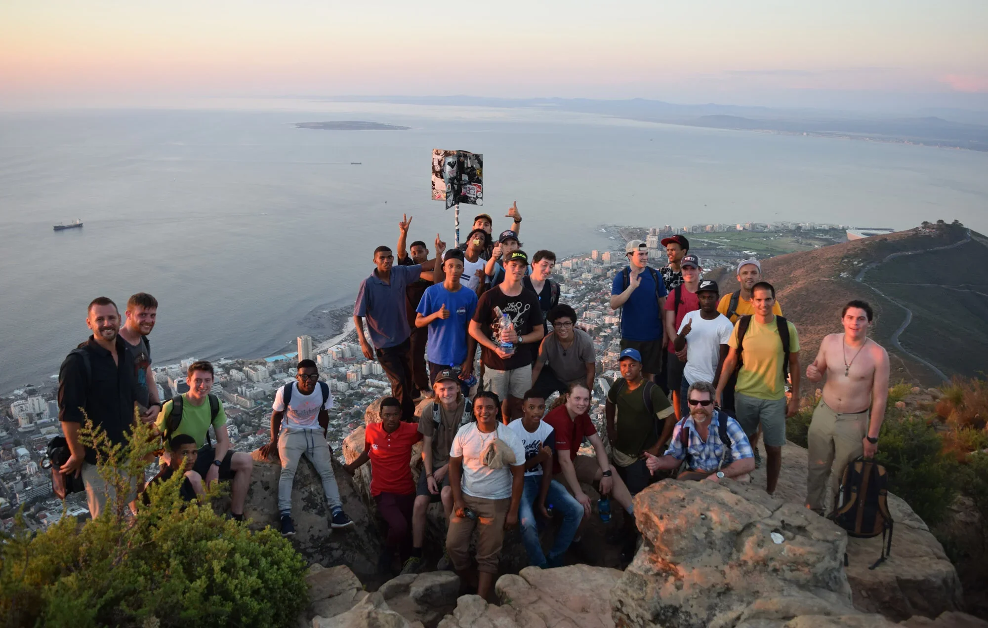 A group of young people and adults posing atop a mountain, with a cityscape and large body of water in the background at sunset.