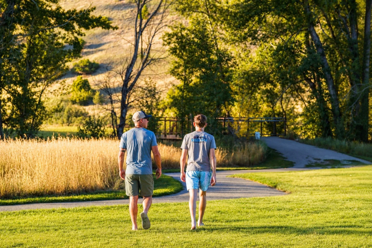 Two people, an adult man and a teenage boy, walk along a park path surrounded by grass and trees during sunset.