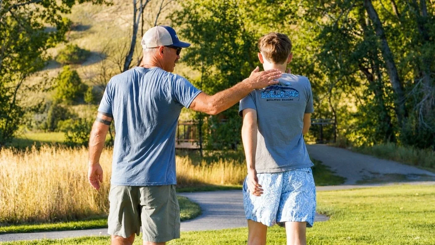 An elderly man with sunglasses and a baseball cap gently touching a young boy's shoulder outdoors in a park during daytime.