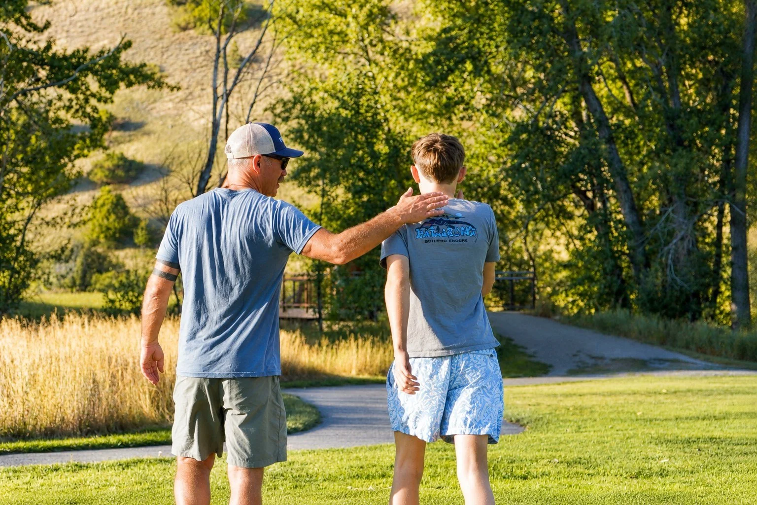 An elderly man and a young boy walk together on a grassy park path during the daytime, surrounded by green trees and nature.