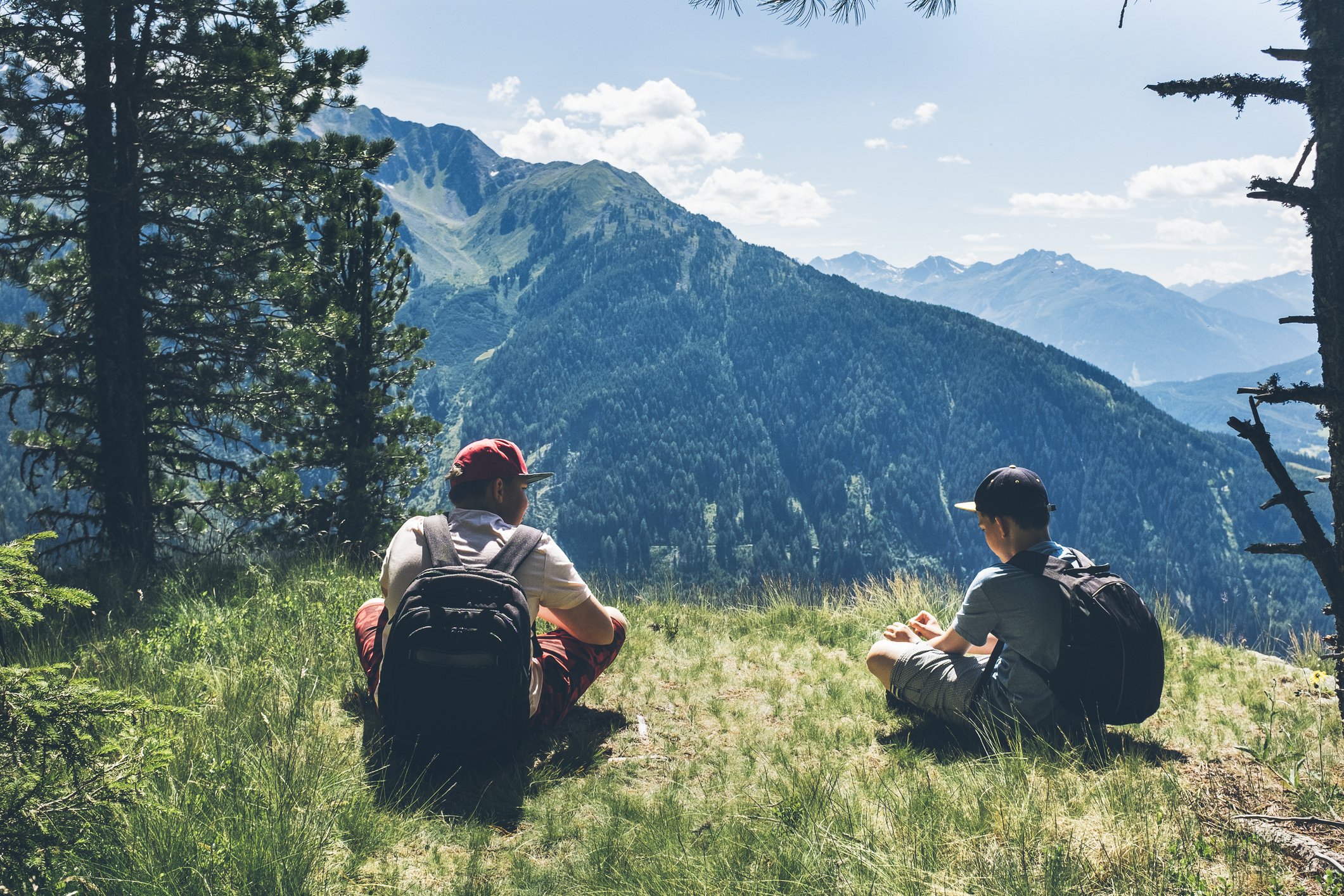 Two hikers with backpacks sitting on grass in a mountain meadow, surrounded by tall pine trees, with a backdrop of rugged mountains and a partly cloudy sky.
