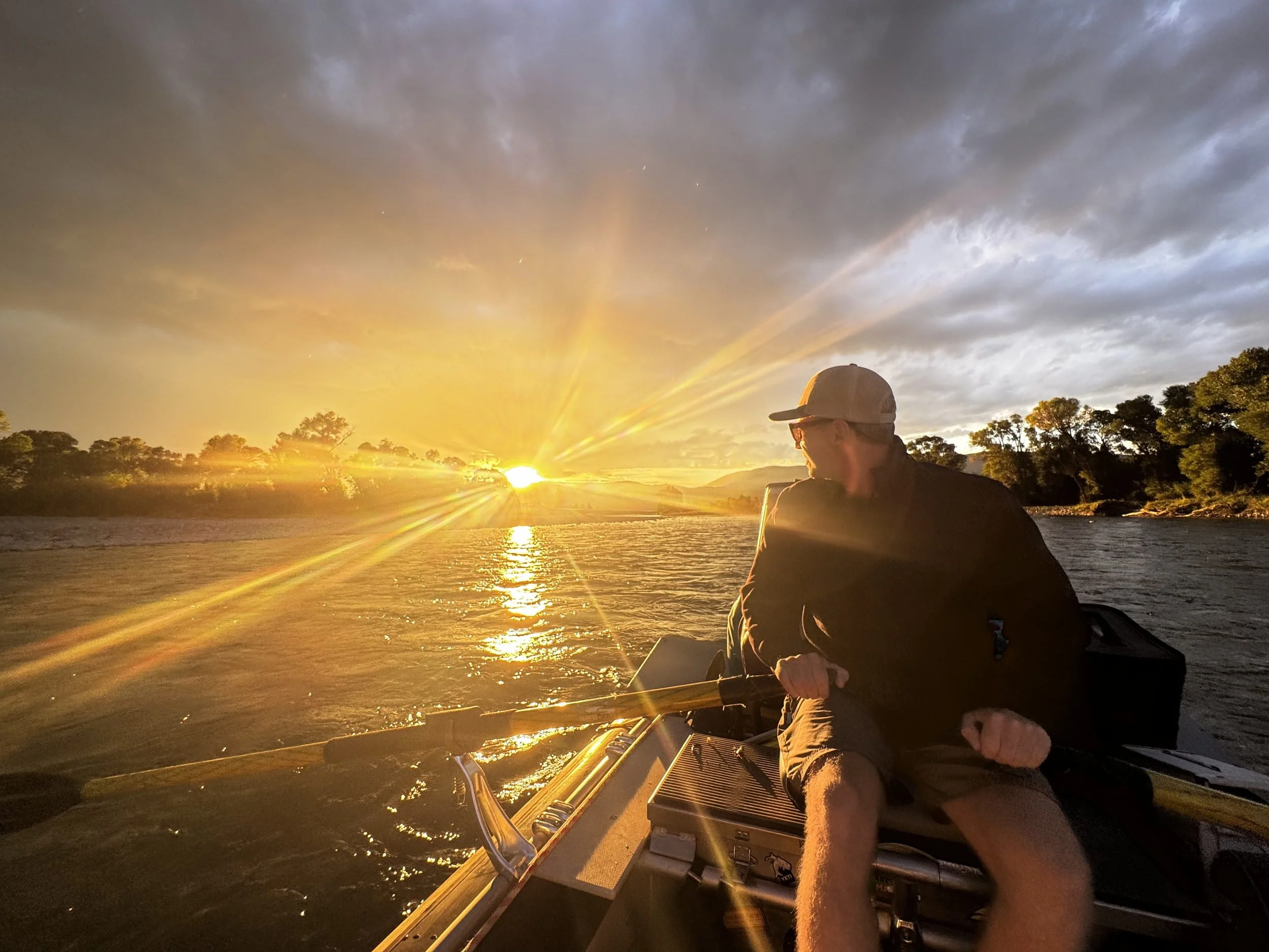 A person wearing a baseball cap and sunglasses sitting in a boat on a river at sunset, with the sun's rays illuminating the sky and reflecting on the water, and trees visible in the background.