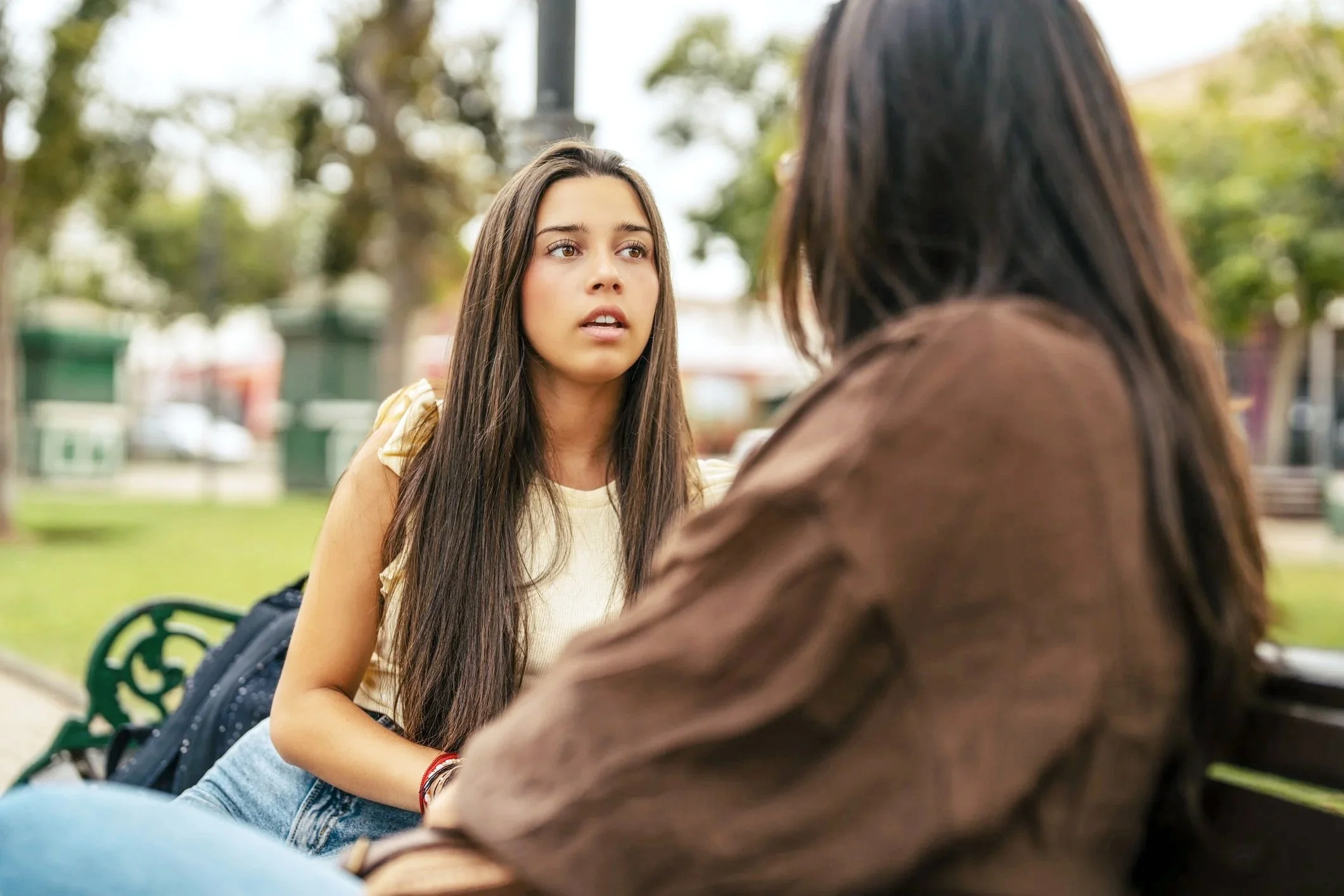 Young woman with long brown hair sitting on a park bench, looking at another person with longues hair, outdoors with trees and park benches in the background.