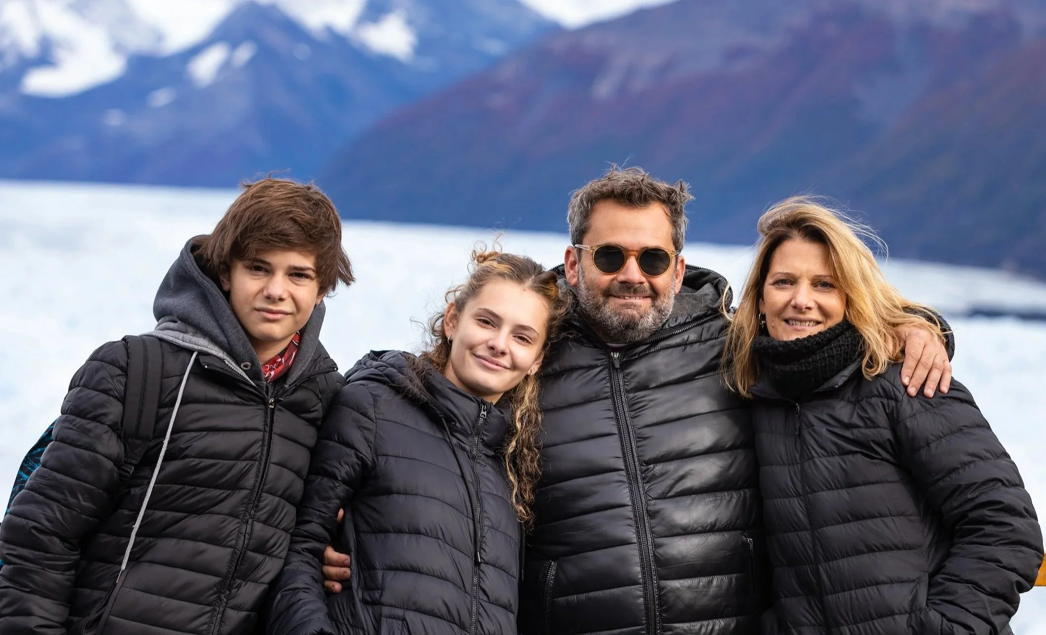 A family of four outdoors, all wearing black jackets, posing with mountains in the background. The father is wearing sunglasses, and the children are smiling.