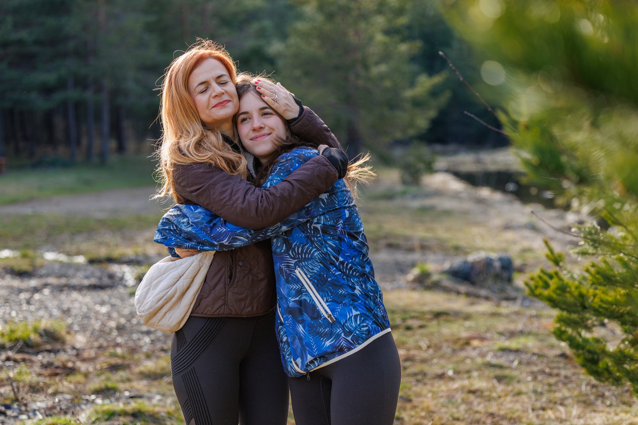 Mother and daughter hugging outdoors in a forested area with sunlight filtering through trees.