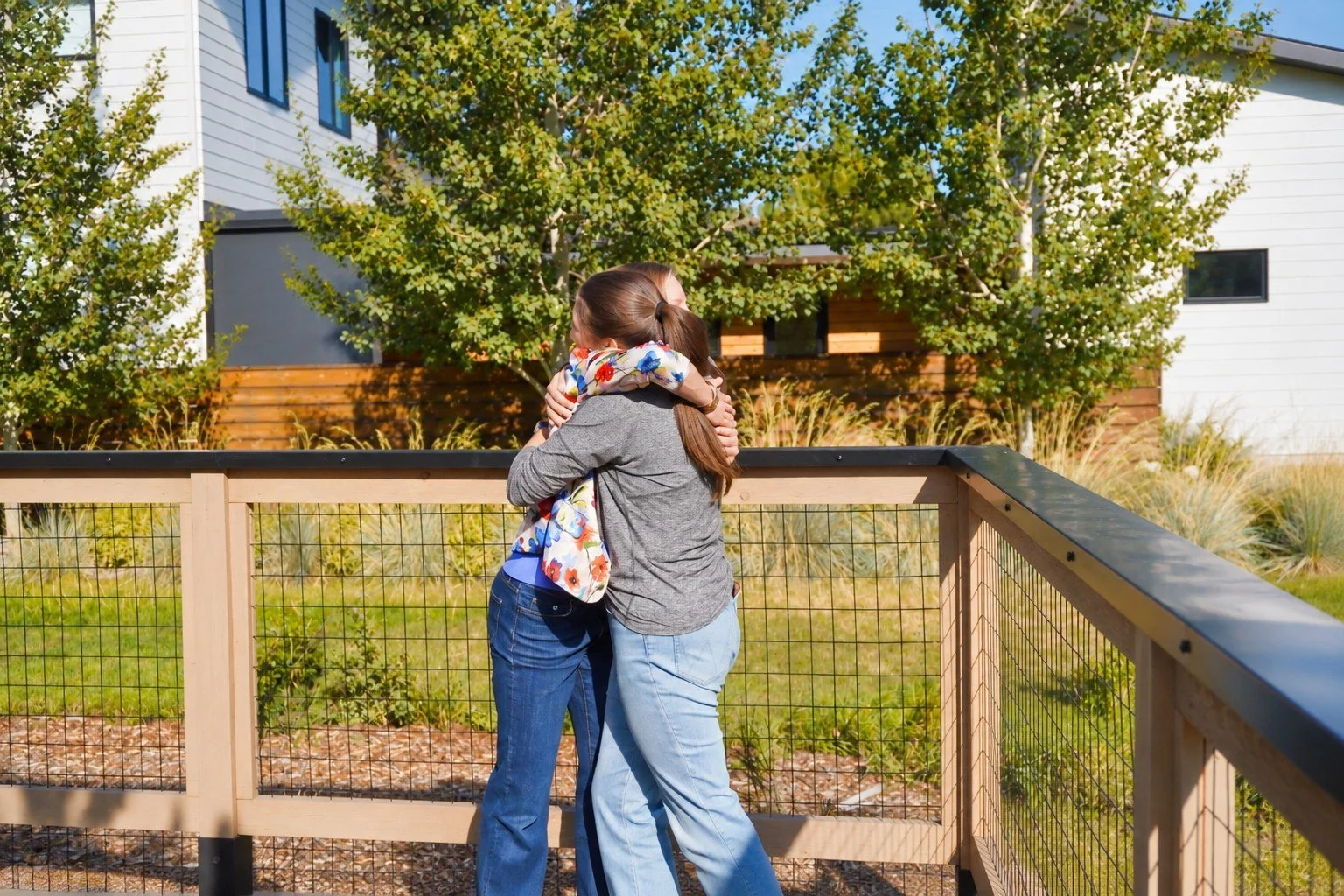 Two women embracing and hugging on a wooden and wire fence outdoor with trees and modern buildings in the background.