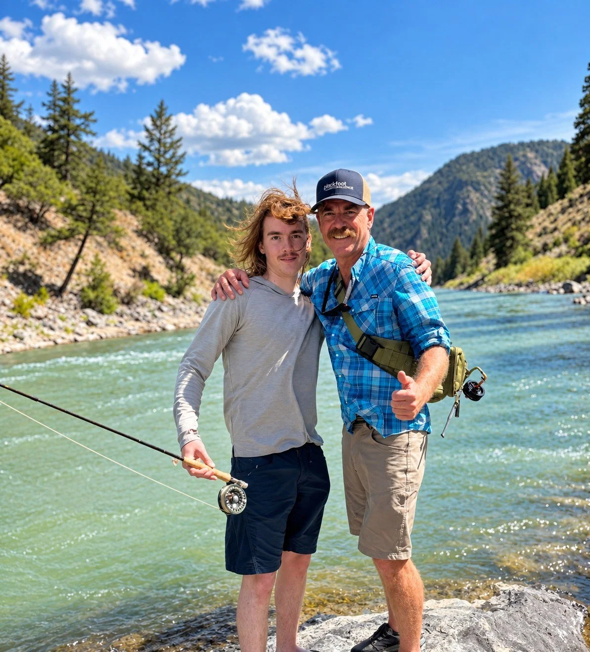 Two men stand close together on rocks by a river, surrounded by mountains and trees under a blue sky with scattered clouds, both smiling and raising their thumbs.