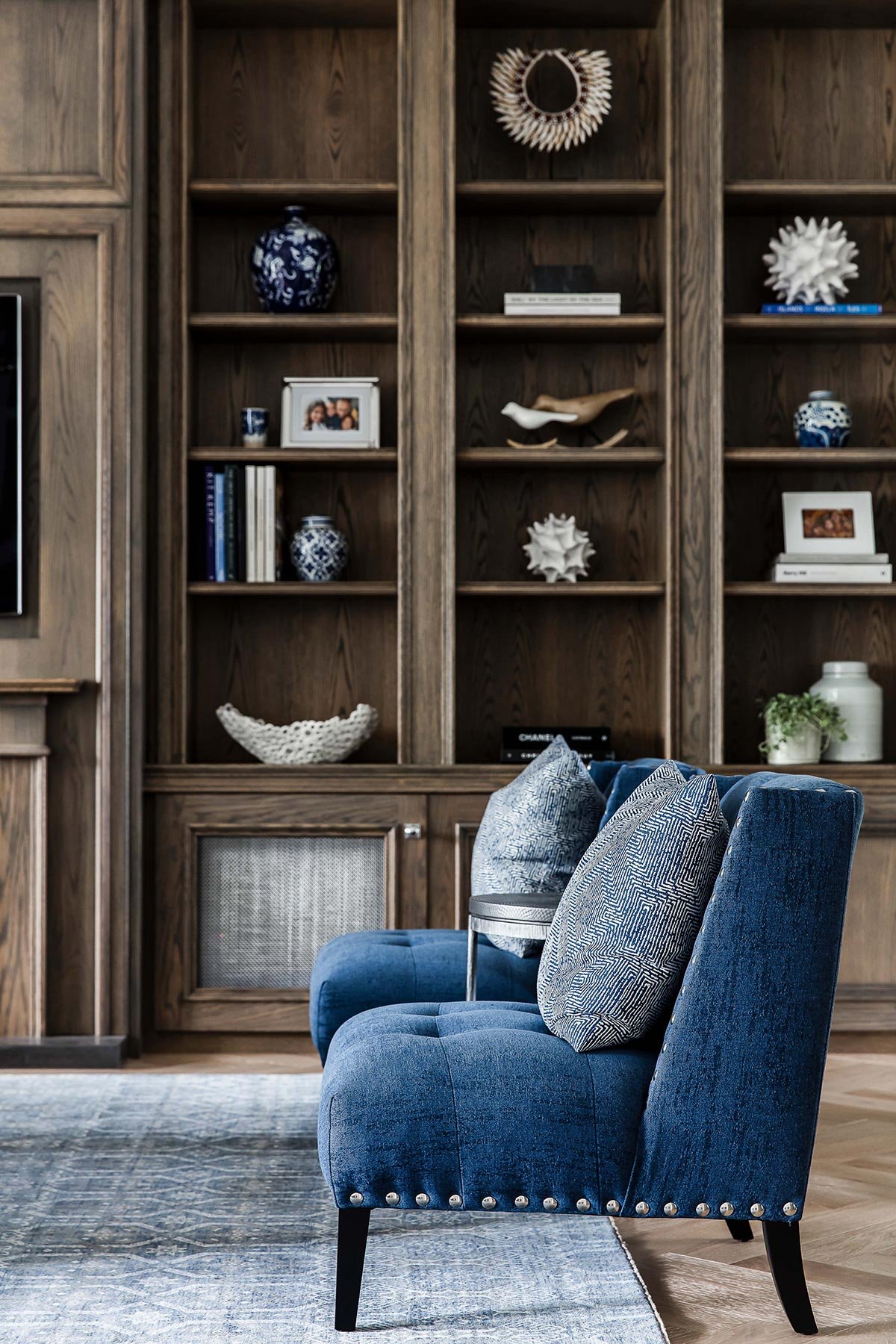 A living room with a wooden bookshelf filled with decorative items, photos, and books in the background. In the foreground, there is a blue velvet chair with patterned pillows on it, placed on a light-colored rug.