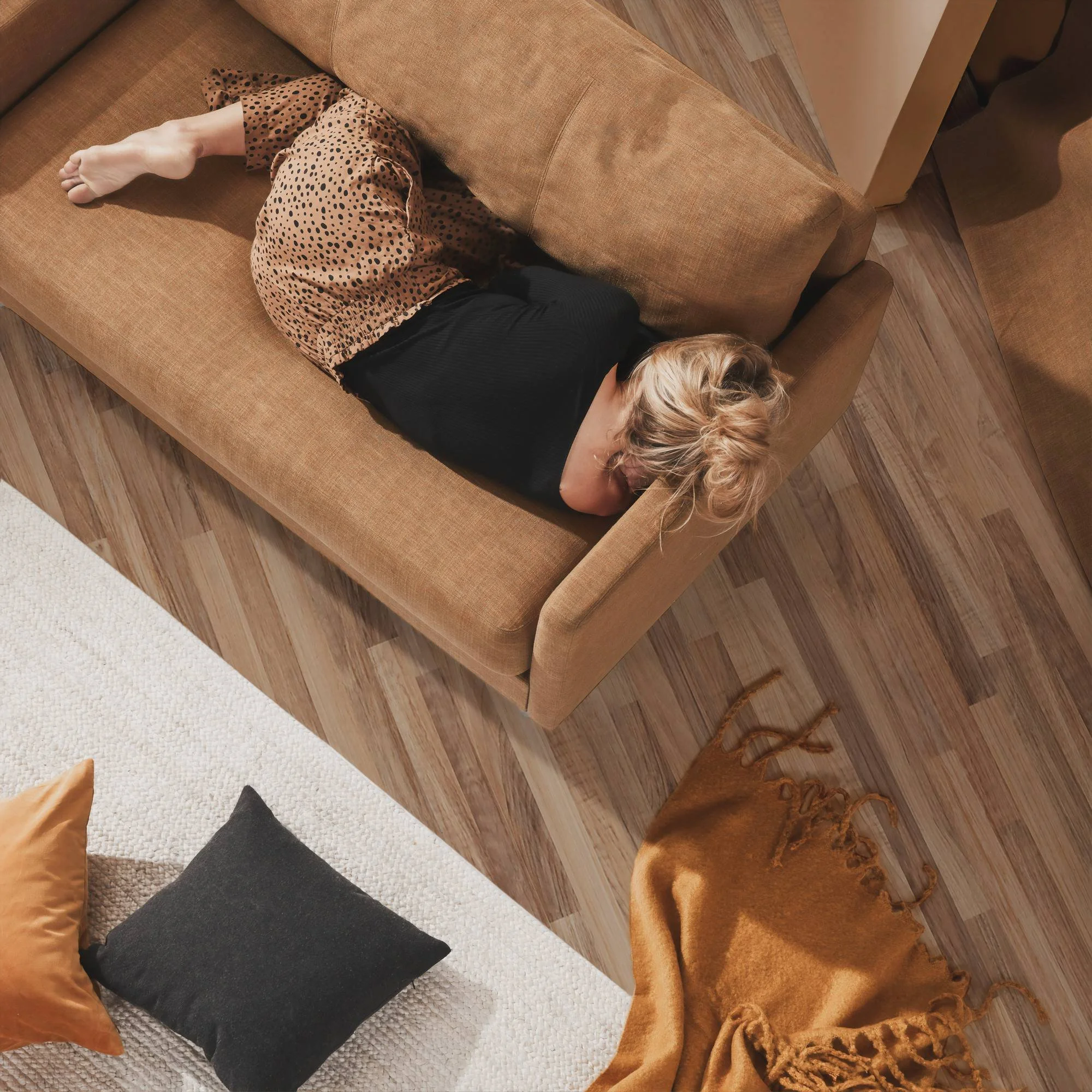 Person lying on a brown upholstered sofa, wearing a polka-dot dress and black leggings, with feet partially covered by the dress, alongside pillows, a blanket, and a wooden floor.