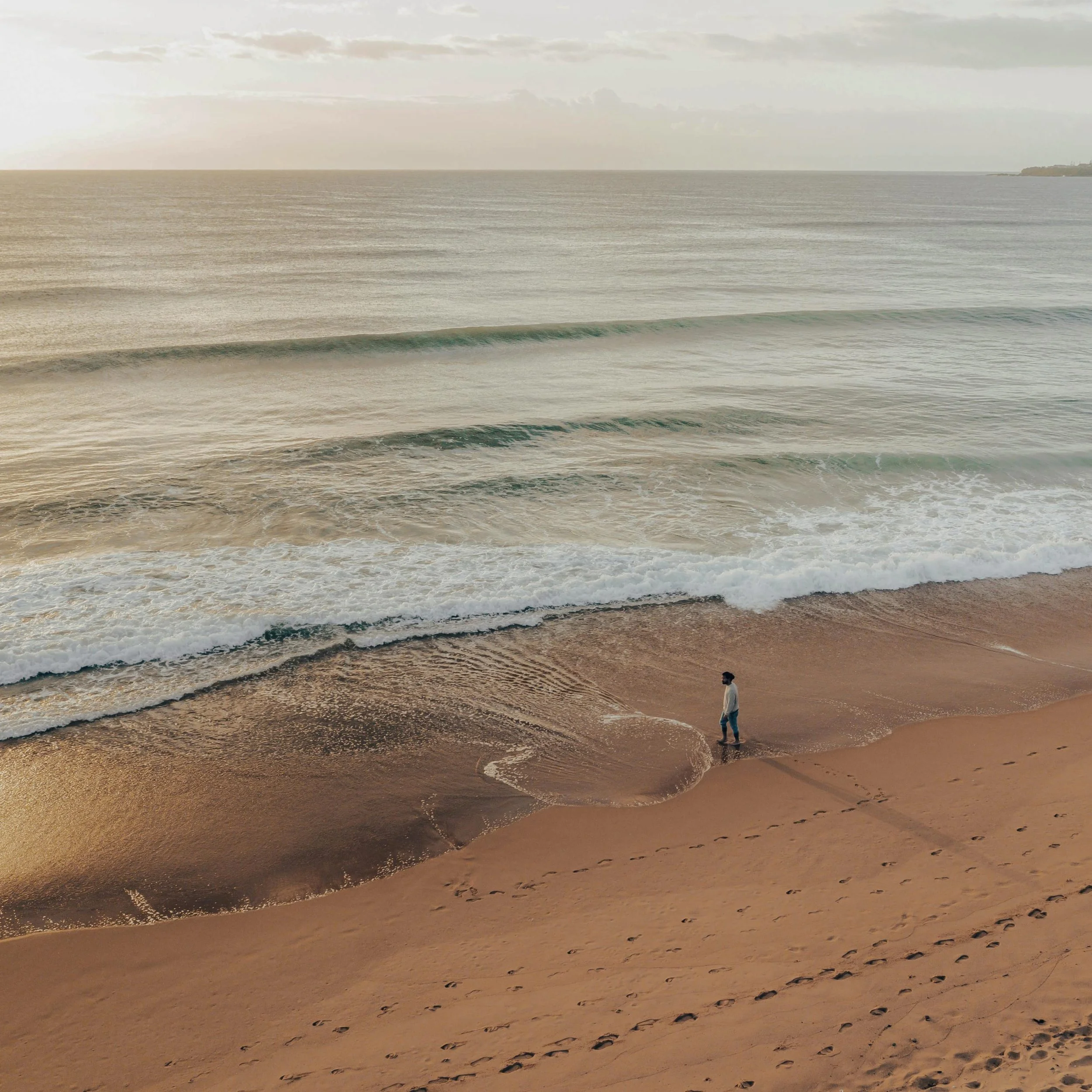 People walking along a sandy beach with footprints, gentle waves, and an overcast sky.