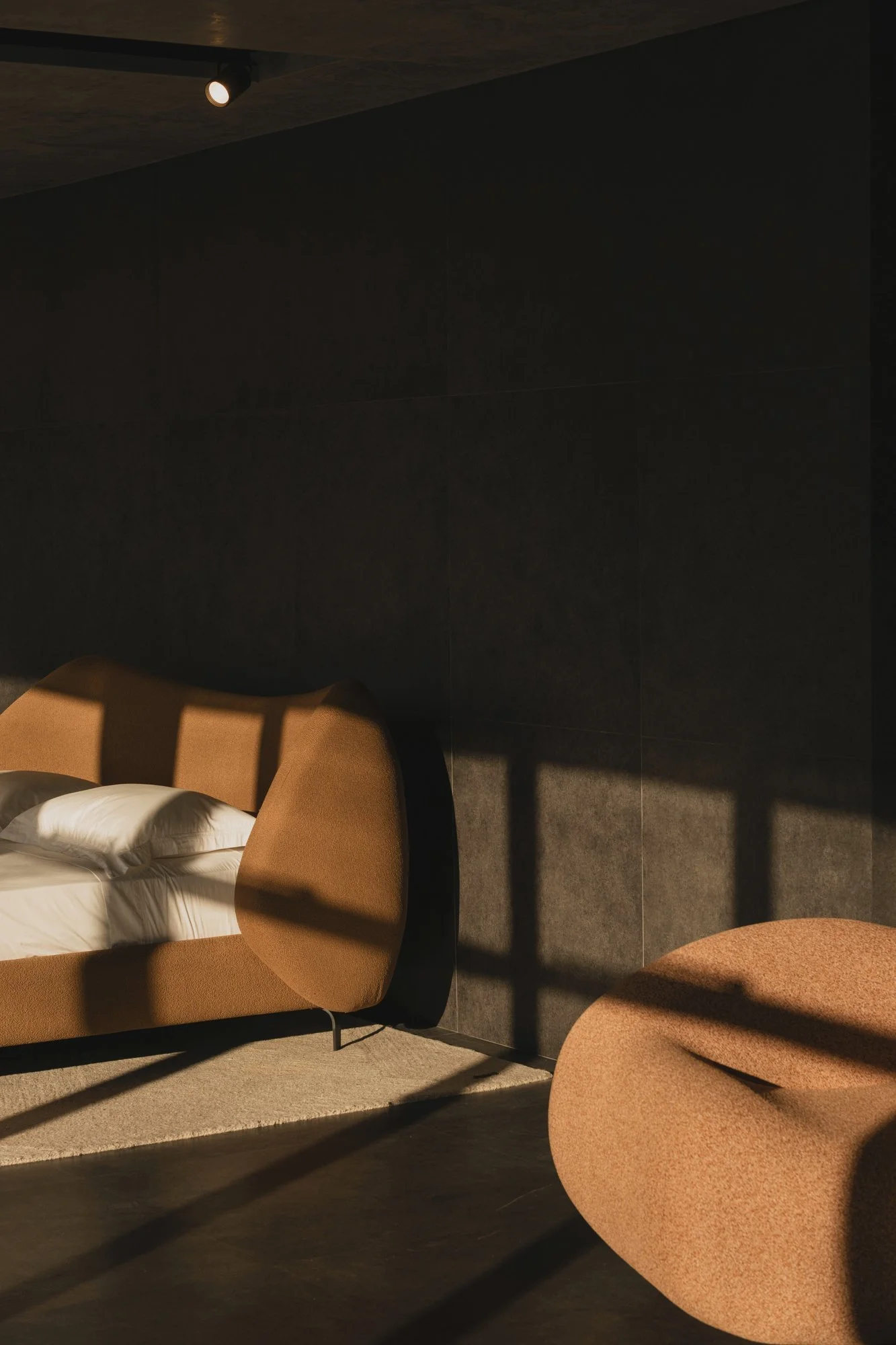 A modern hotel room featuring a bed with white linens, a tan upholstered armchair, and a matching round ottoman, with black walls and shadows cast by window blinds.