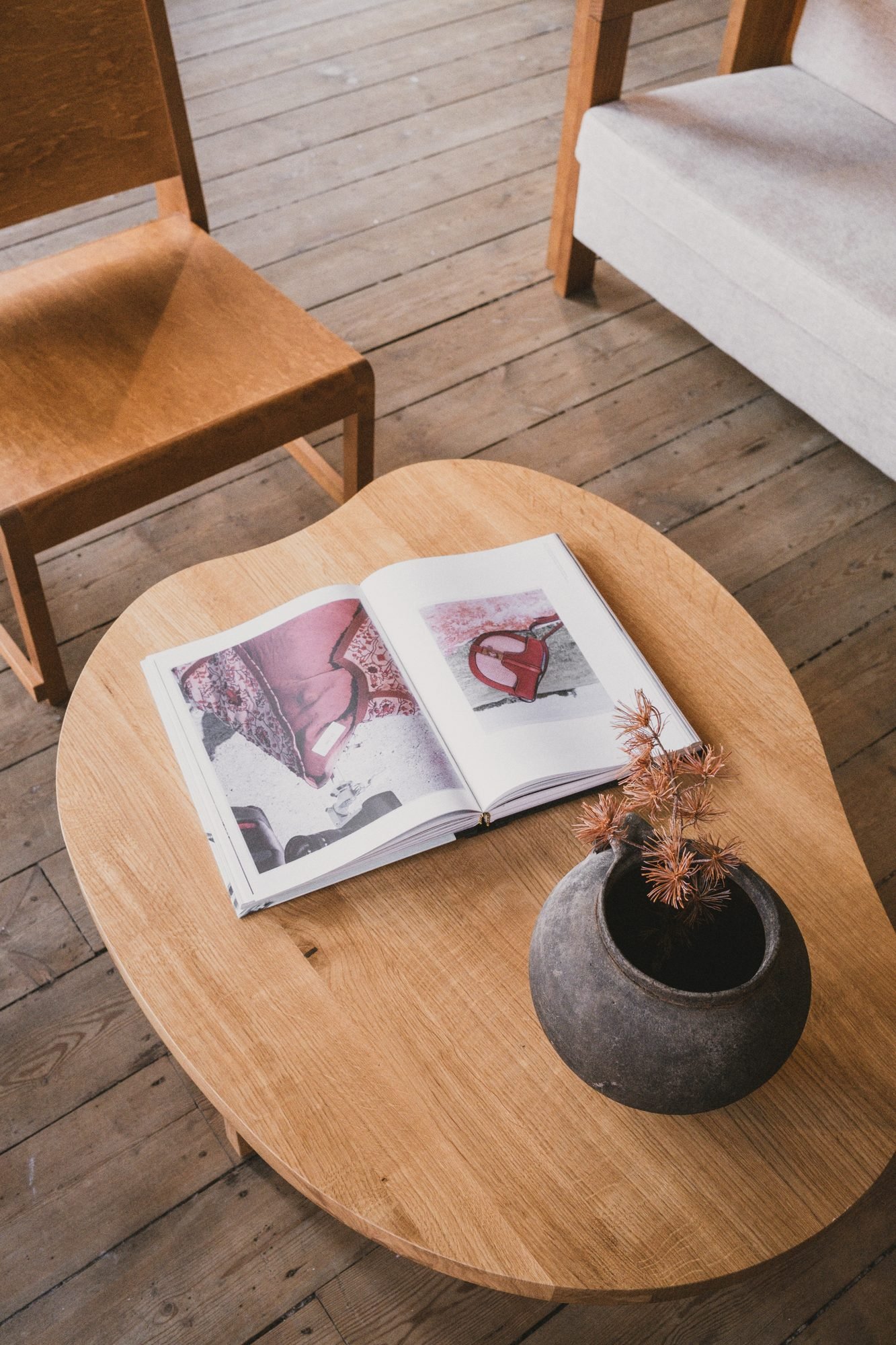A wooden coffee table with an open magazine showing images of handbags, alongside a decorative vase with dried flowers inside.