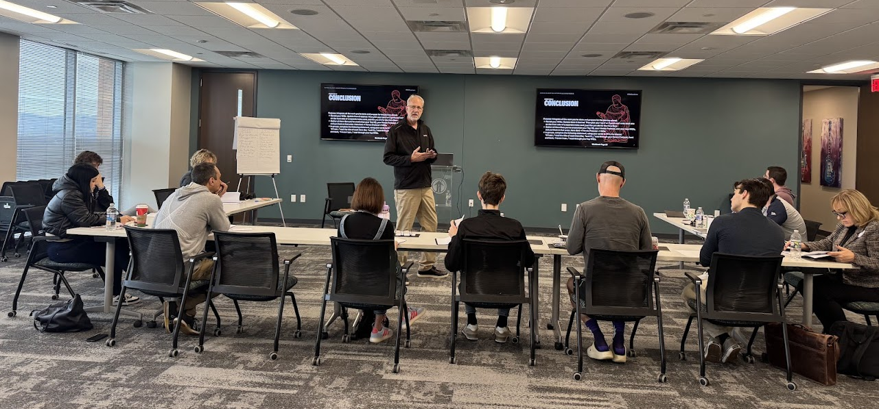 A man giving a presentation in a conference room with attendees seated at tables listening. Two screens display the conclusion of his presentation.