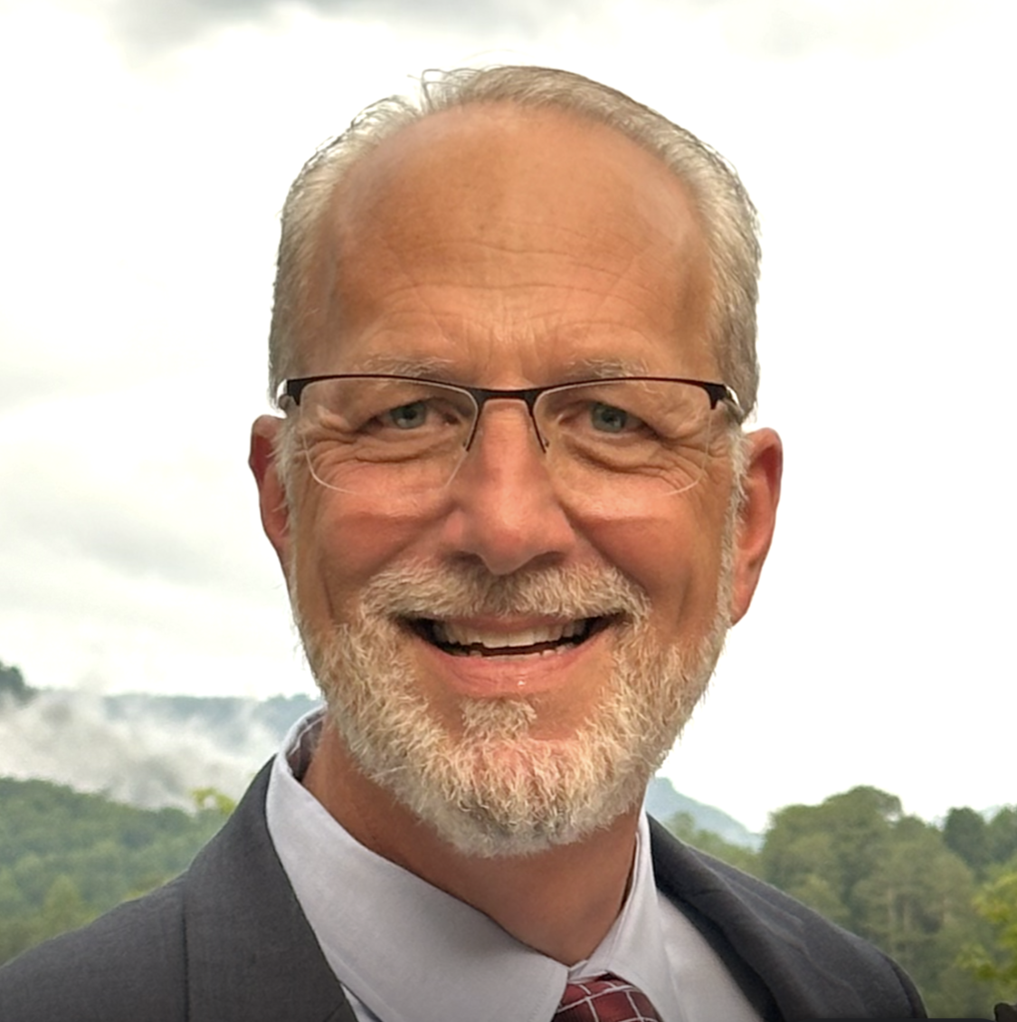 A middle-aged man with glasses and a beard smiling outdoors with a cloudy sky and distant hills in the background.  Curt Hollmann.
