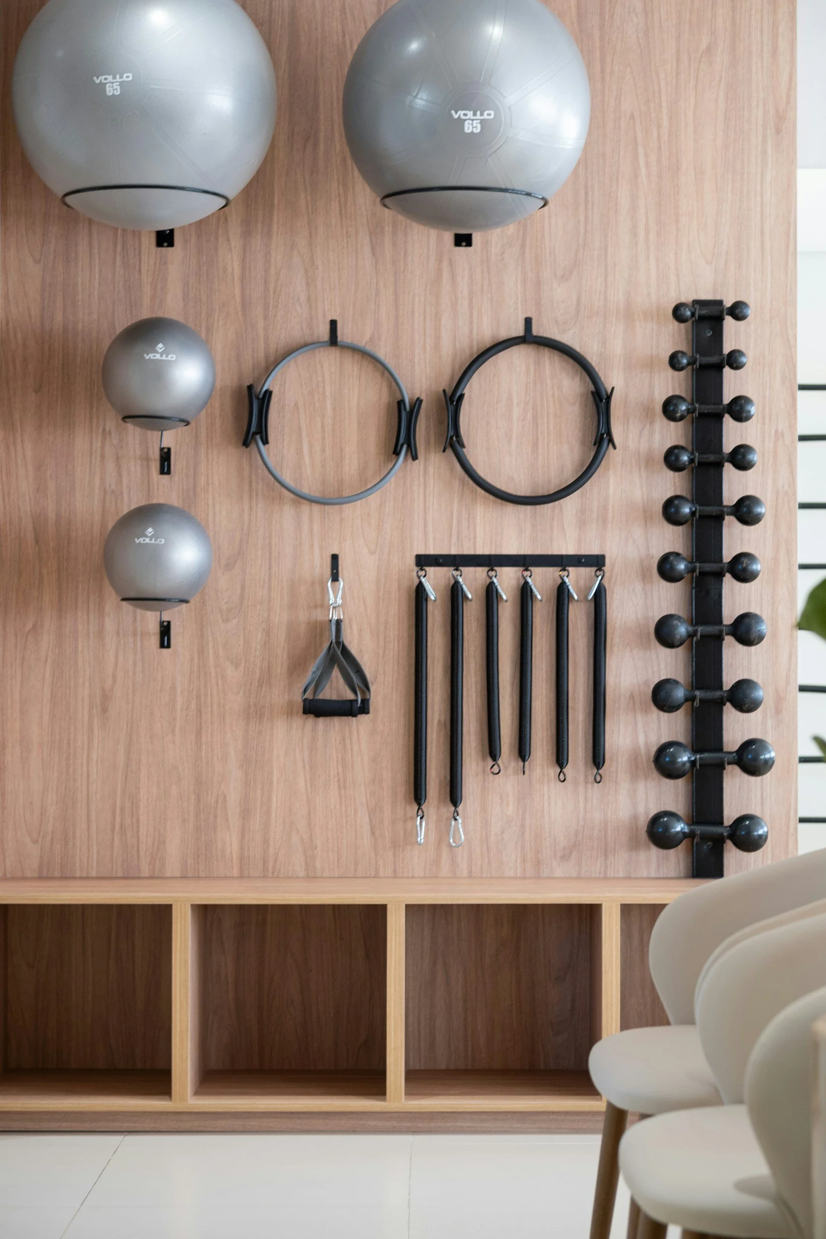 Workout equipment wall with hanging exercise balls, resistance rings, bands, and a rack of small weights. Chairs are visible in the foreground.