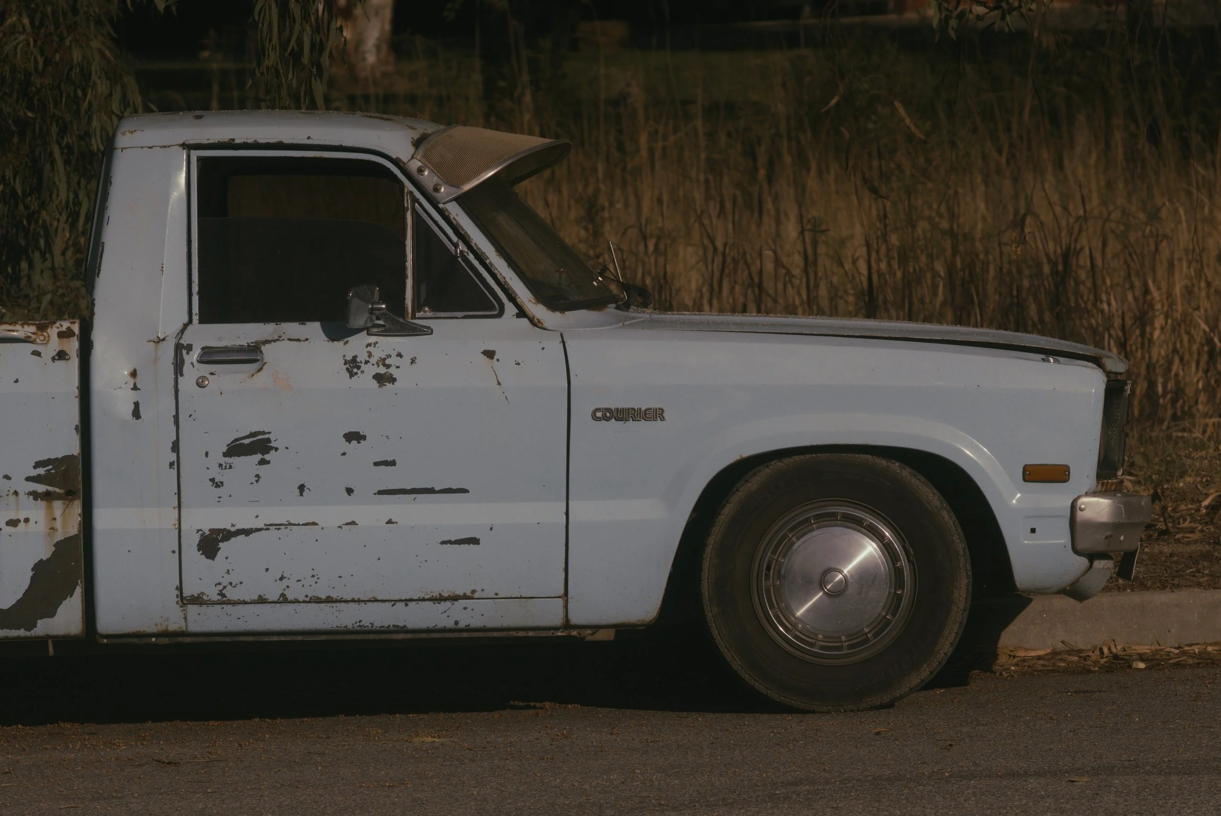 A vintage white pickup truck with rust and wear marks parked on the side of the road at night, with dry grass and trees in the background.