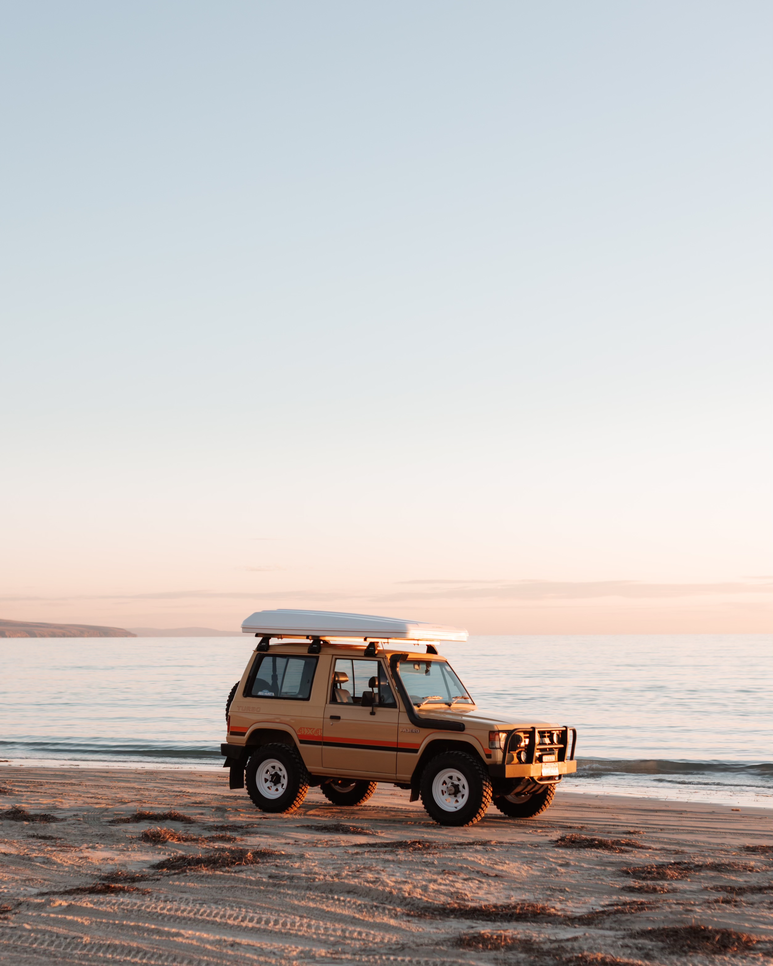 A beige off-road vehicle with a white roof parked on a sandy beach at sunset, with calm water and a distant shoreline in the background.