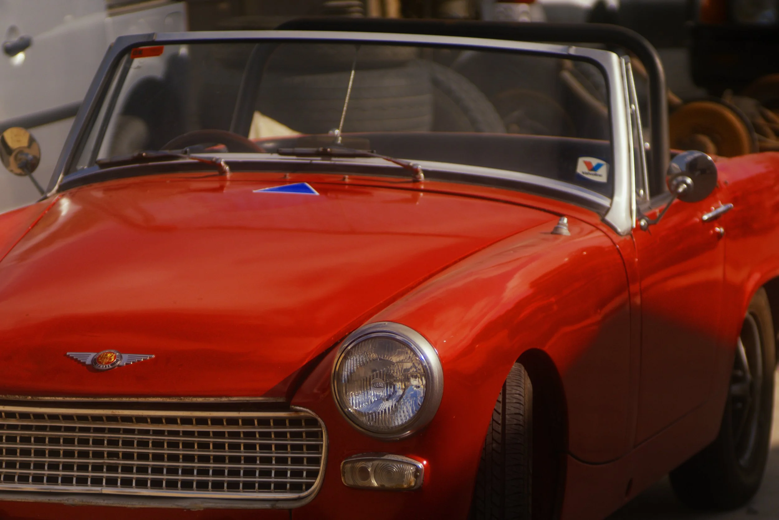 Close-up of a vintage red sports car, showing the front and part of the driver's side, including the headlights, grill, and windshield.