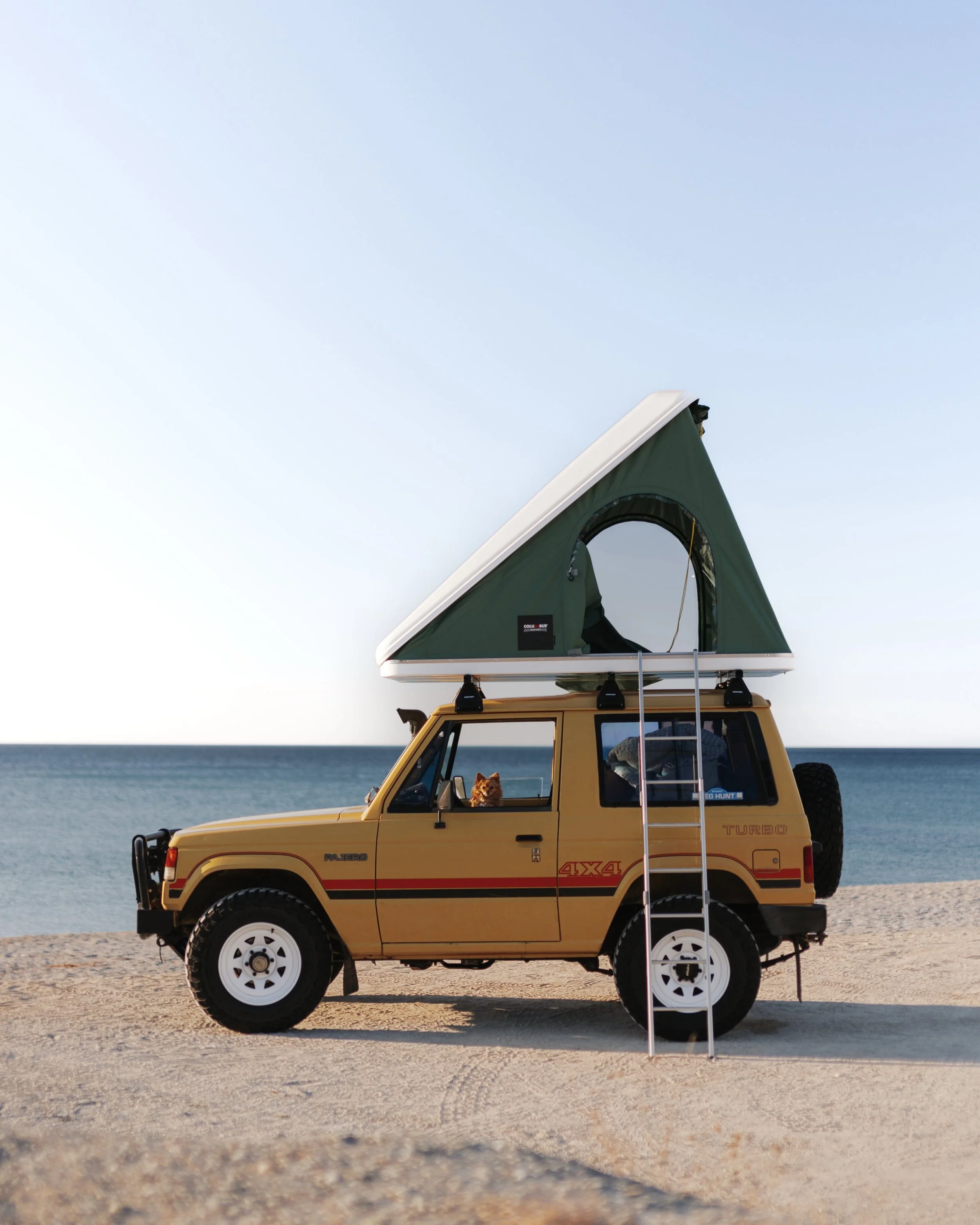 A yellow off-road vehicle with a roof tent mounted on top, parked on a sandy beach near the ocean, with a dog sitting inside and looking out the window.