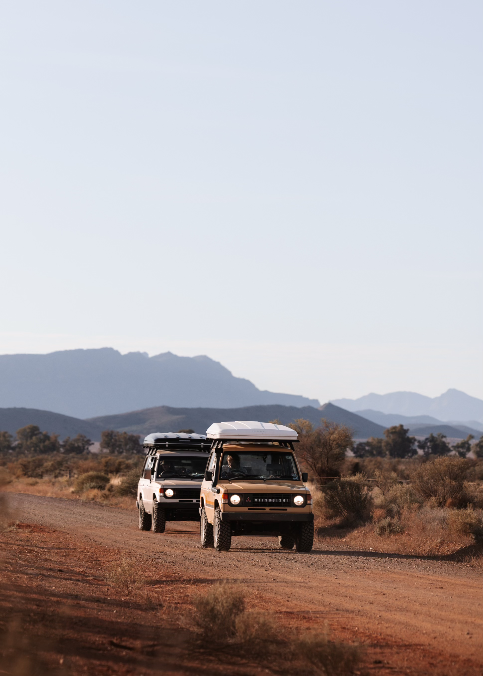 Two off-road SUVs driving on a dirt road in a desert landscape, with mountains in the background and clear sky overhead.