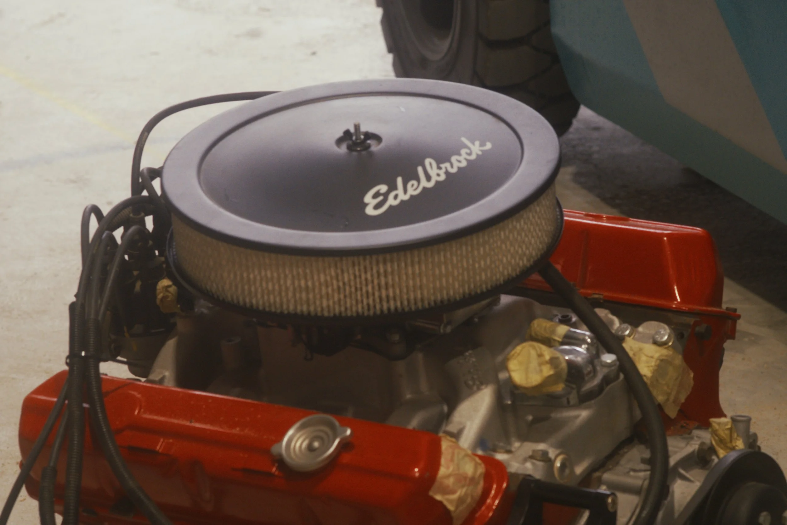 Close-up of a car engine with an Edelbrock logo on the air filter, mounted on a red chassis, with various hoses and parts surrounding it.