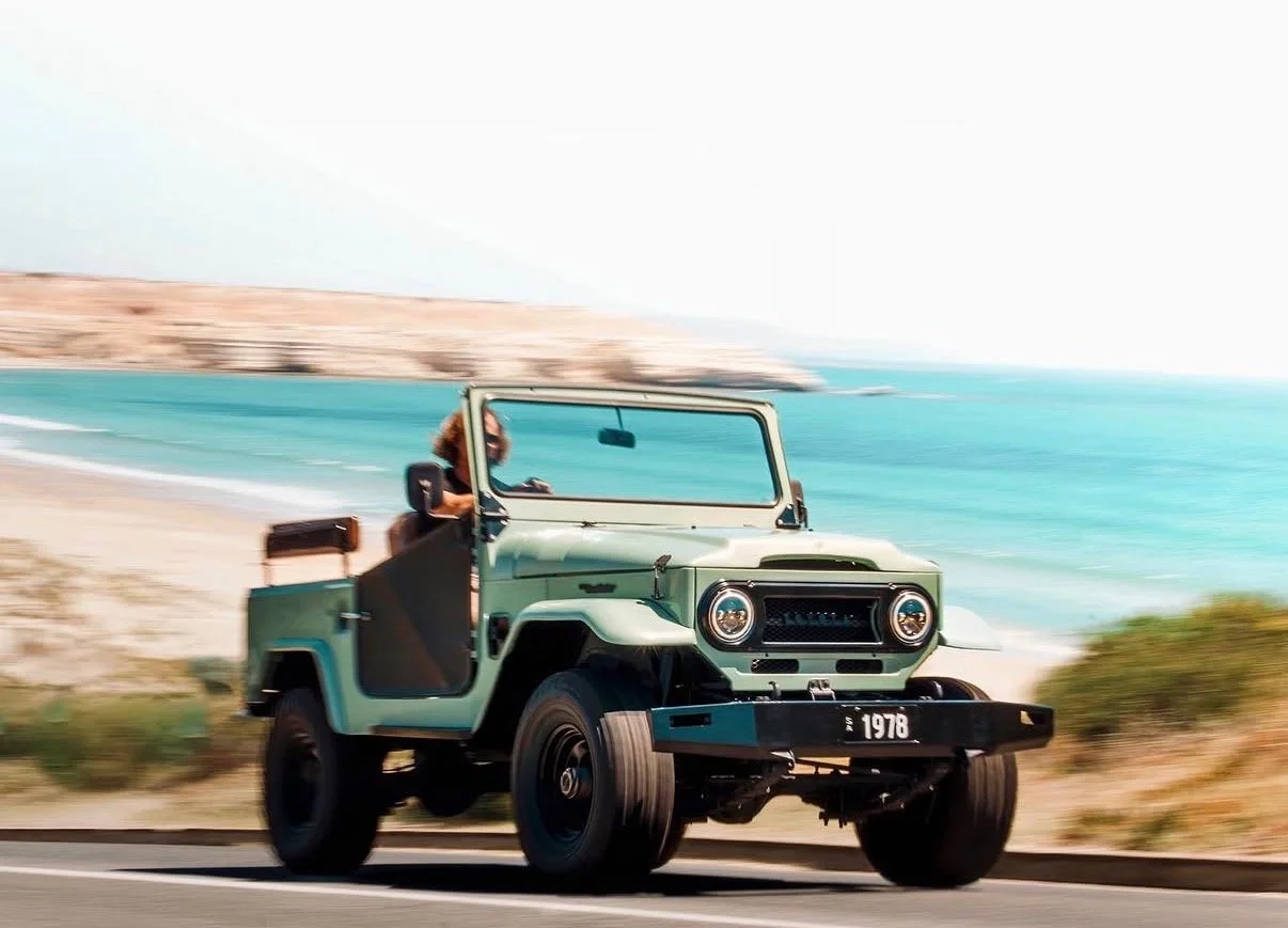 A vintage green off-road vehicle driving along a road by the coast with a woman inside, and the ocean and sandy landscape in the background.
