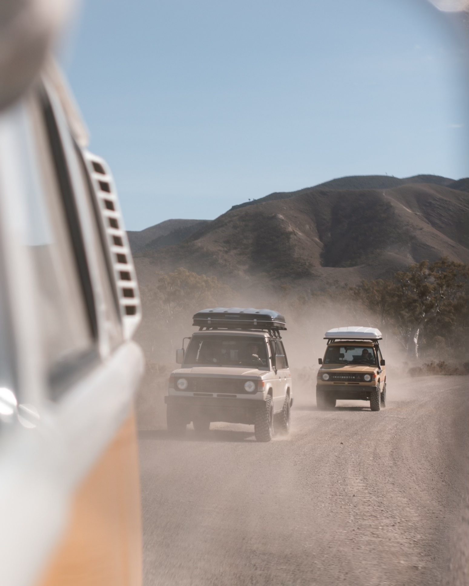Off-road vehicles driving on a dusty dirt road in a desert area with hills in the background, viewed from the side of a larger vehicle.