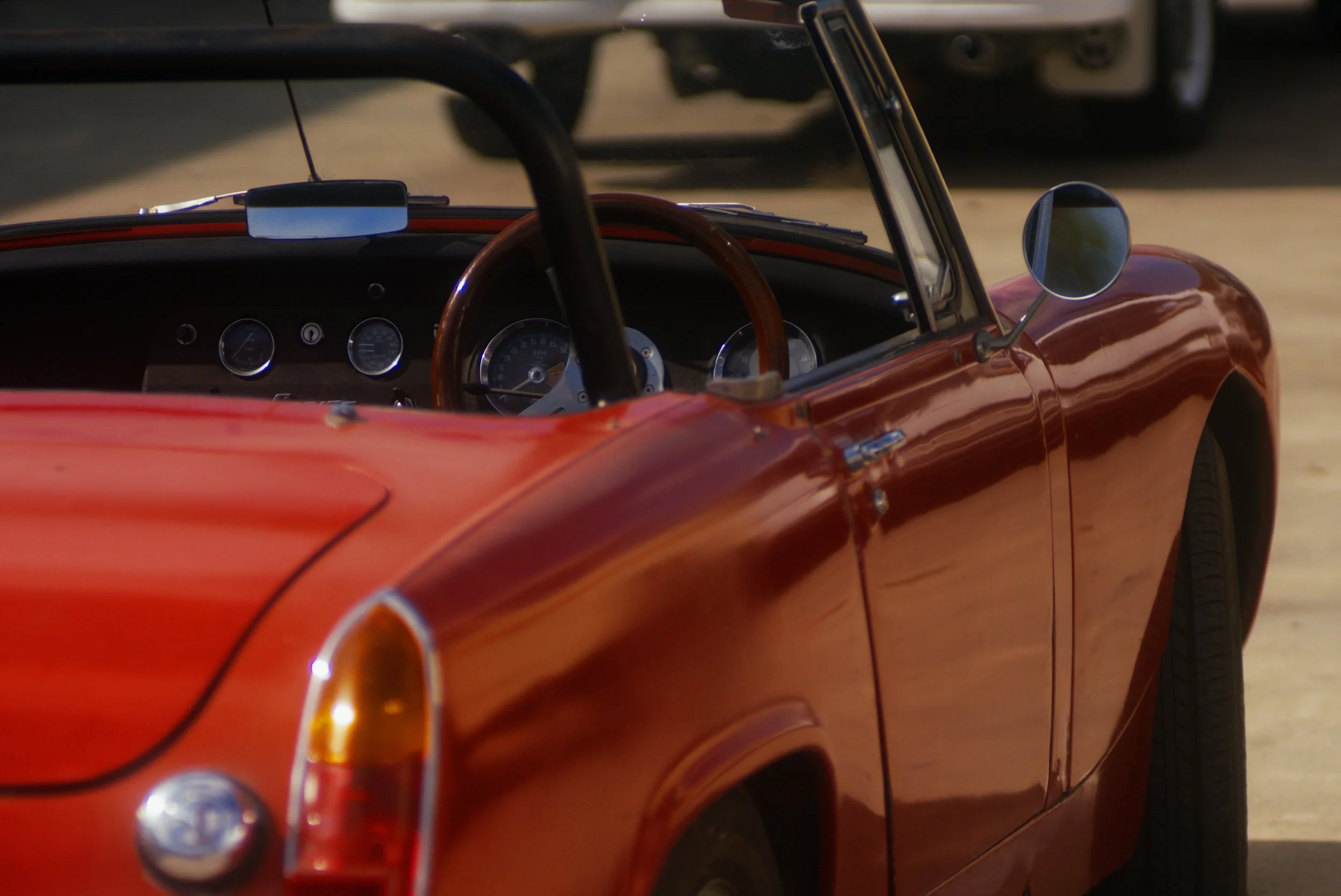 Close-up of a vintage red sports car's dashboard and steering wheel, with the car's side mirror and part of the exterior body visible, set outdoors on a dirt or gravel surface.