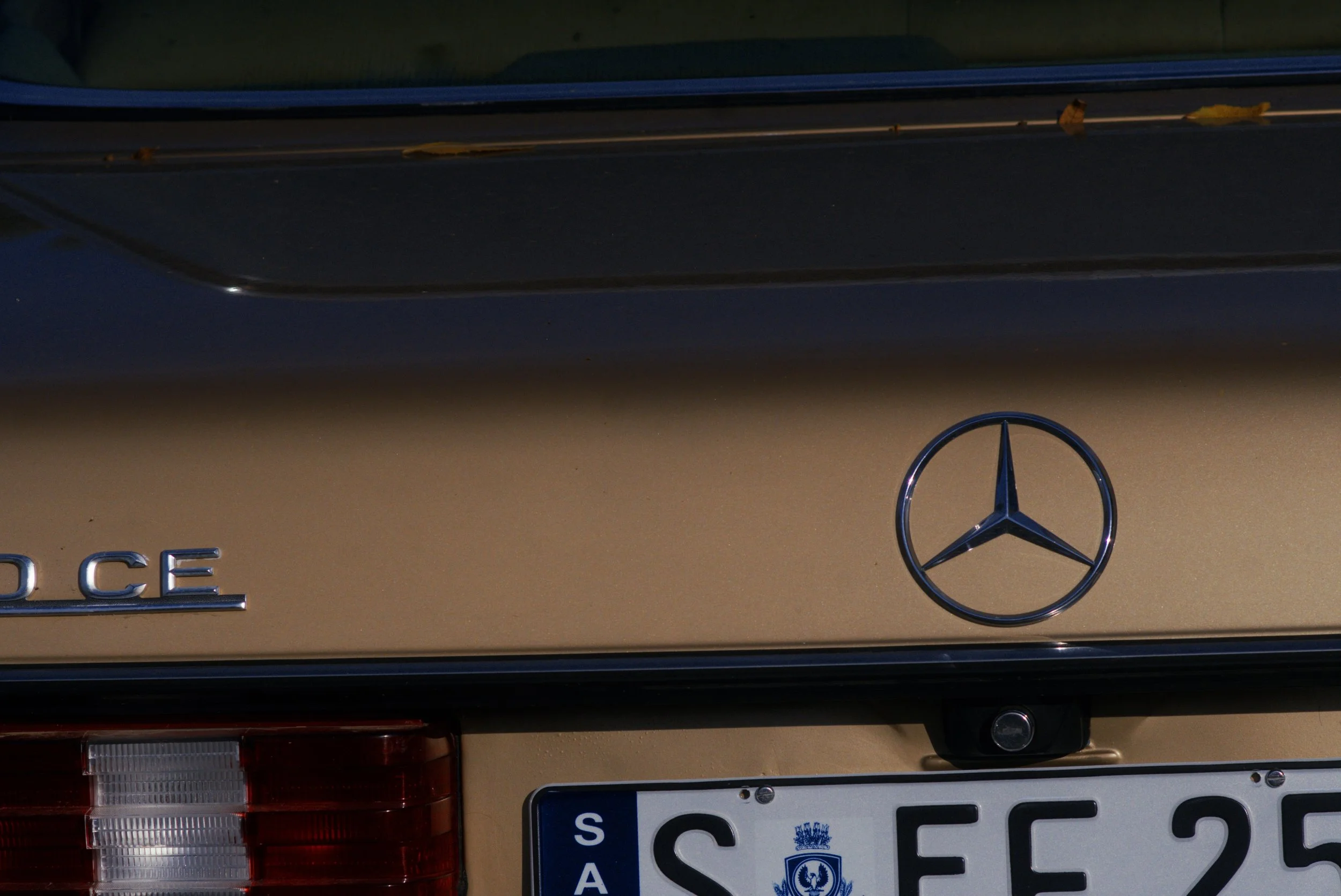 Close-up of the rear of a gold Mercedes-Benz vehicle, showing the Mercedes logo, part of the model name, and a German license plate.