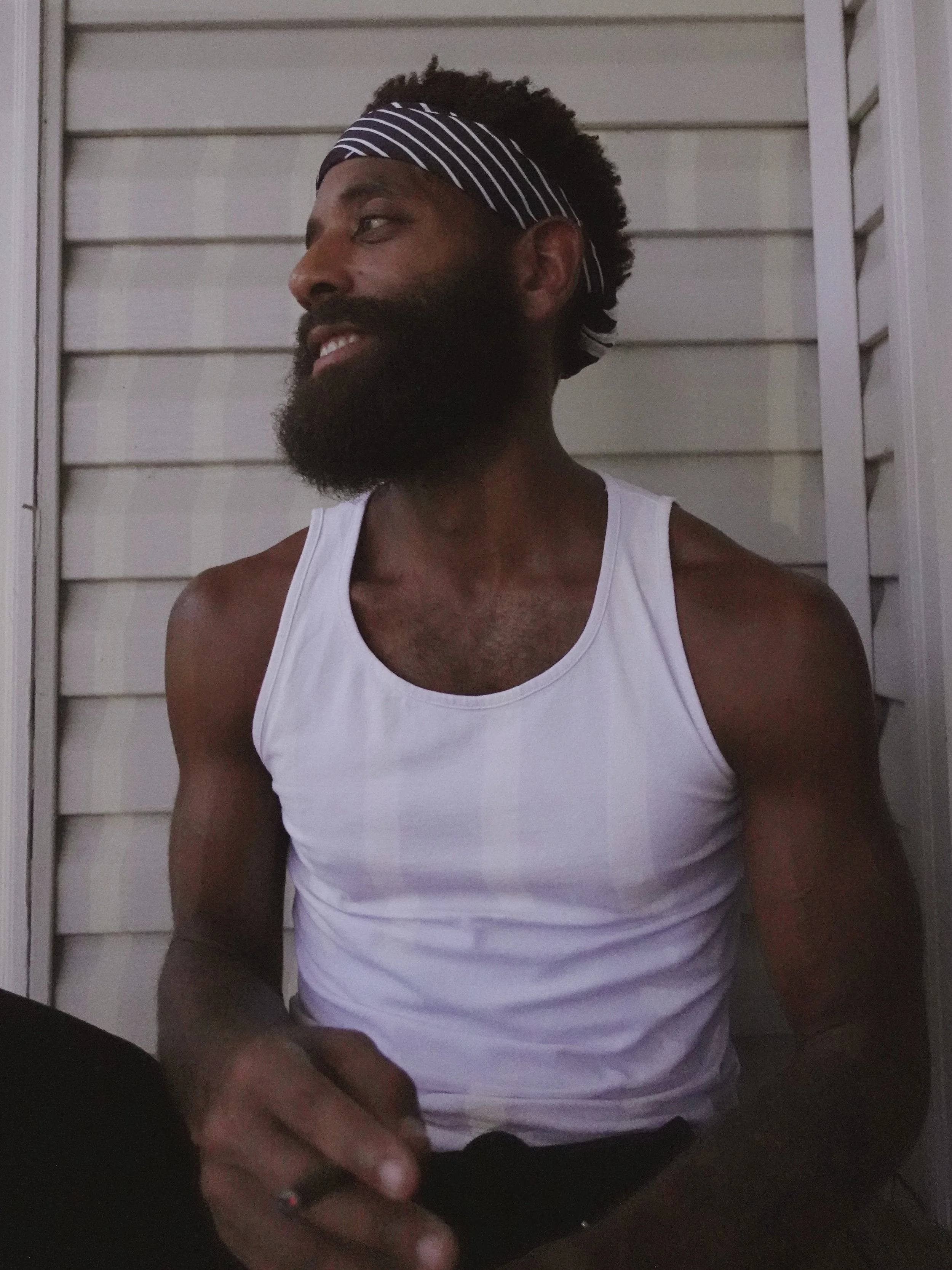 A man with a beard and mustache wearing a striped headband and a white tank top, standing against a beige wooden wall, looking to his left, smiling.
