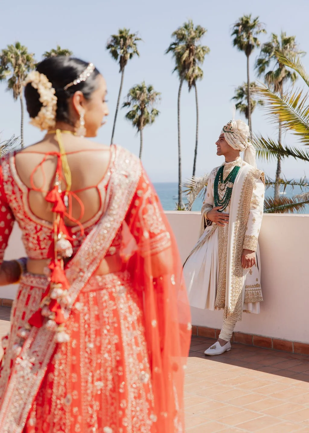 A bride in traditional Indian wedding attire, facing a groom in white and gold traditional outfit, with palm trees and a blue sky in the background.