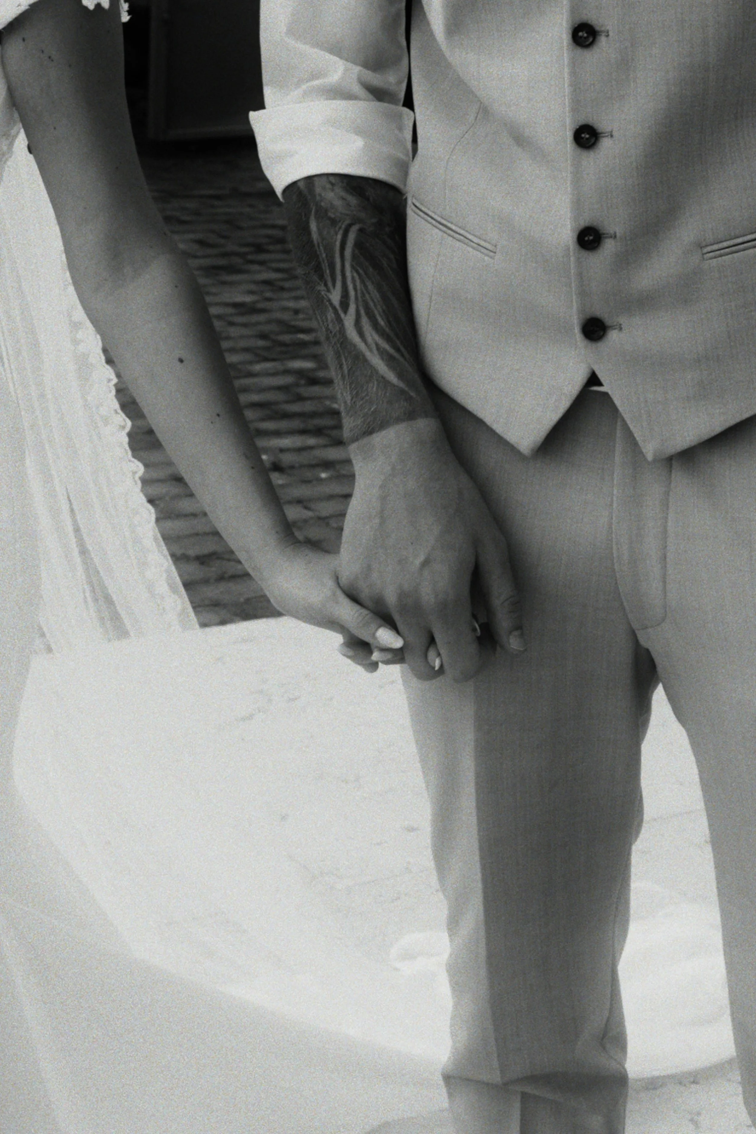 Bride and groom in a light-colored suit holding hands with another, with only their hands, arms, and part of their torso visible.