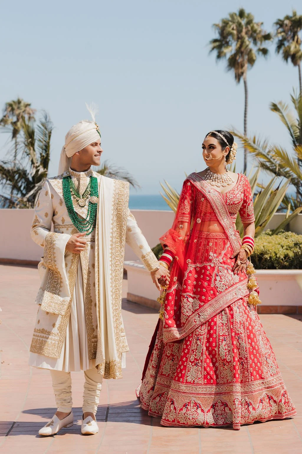 A newly married Indian couple dressed in traditional wedding attire, holding hands outdoors with palm trees, a clear sky, and an ocean in the background.