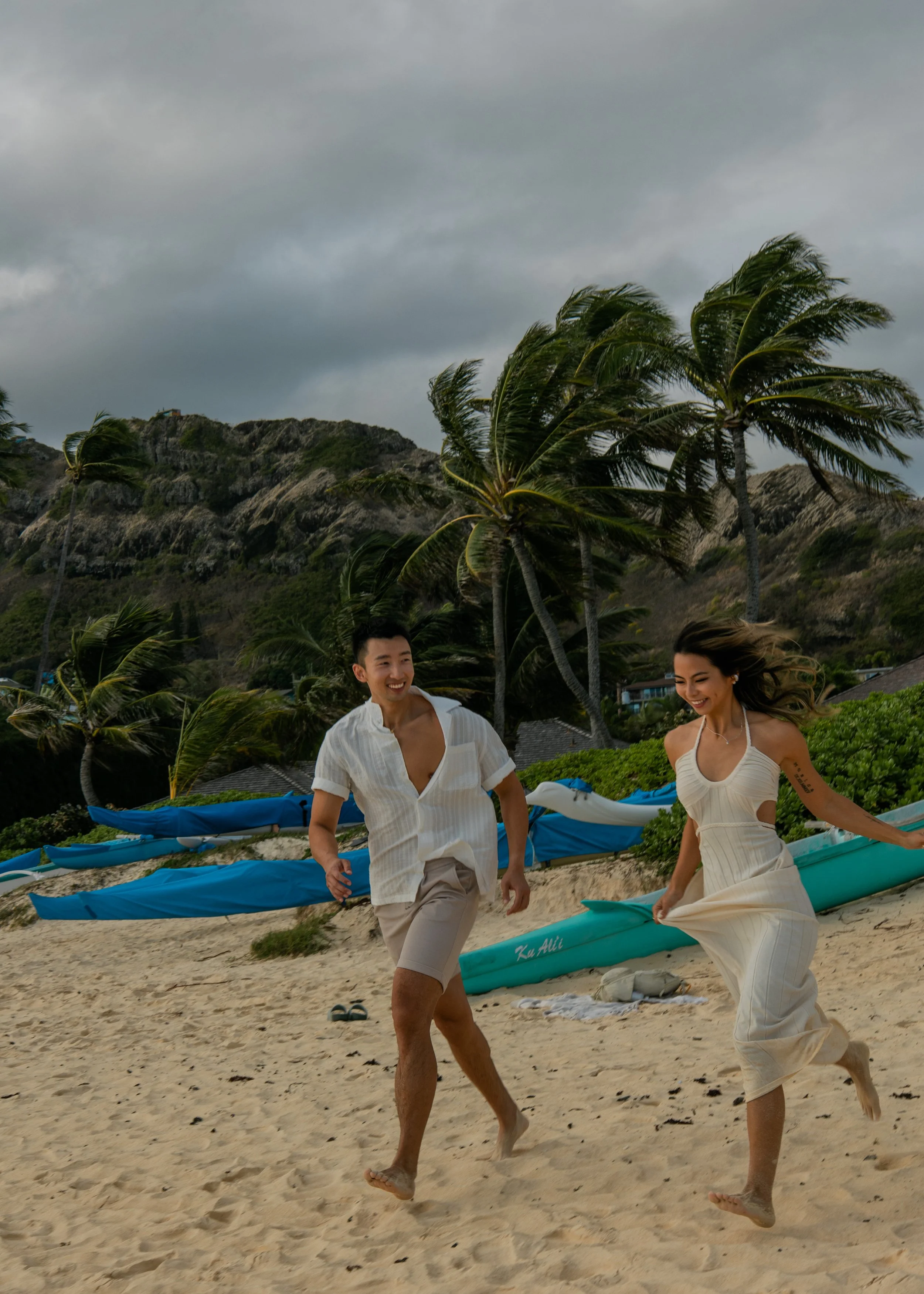 A smiling man and woman running on the beach with palm trees and mountains in the background, cloudy sky overhead, and kayaks on the sand.