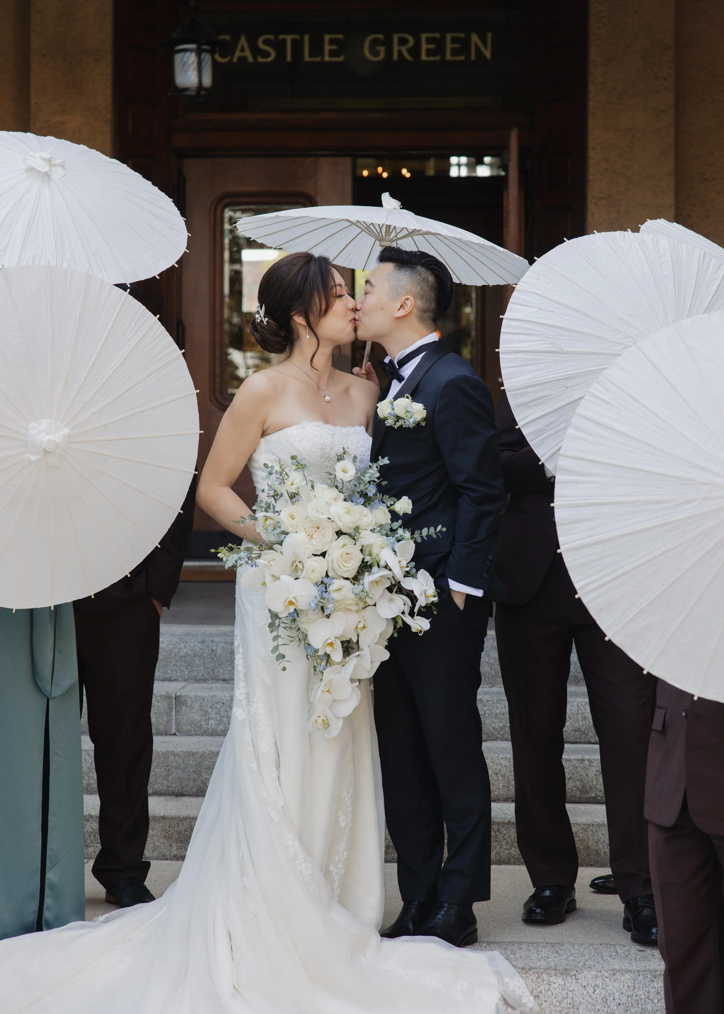 A bride and groom are kissing on steps outside a building labeled 'Castel Green', surrounded by four people holding white paper parasols, with the bride holding a large bouquet of white flowers.