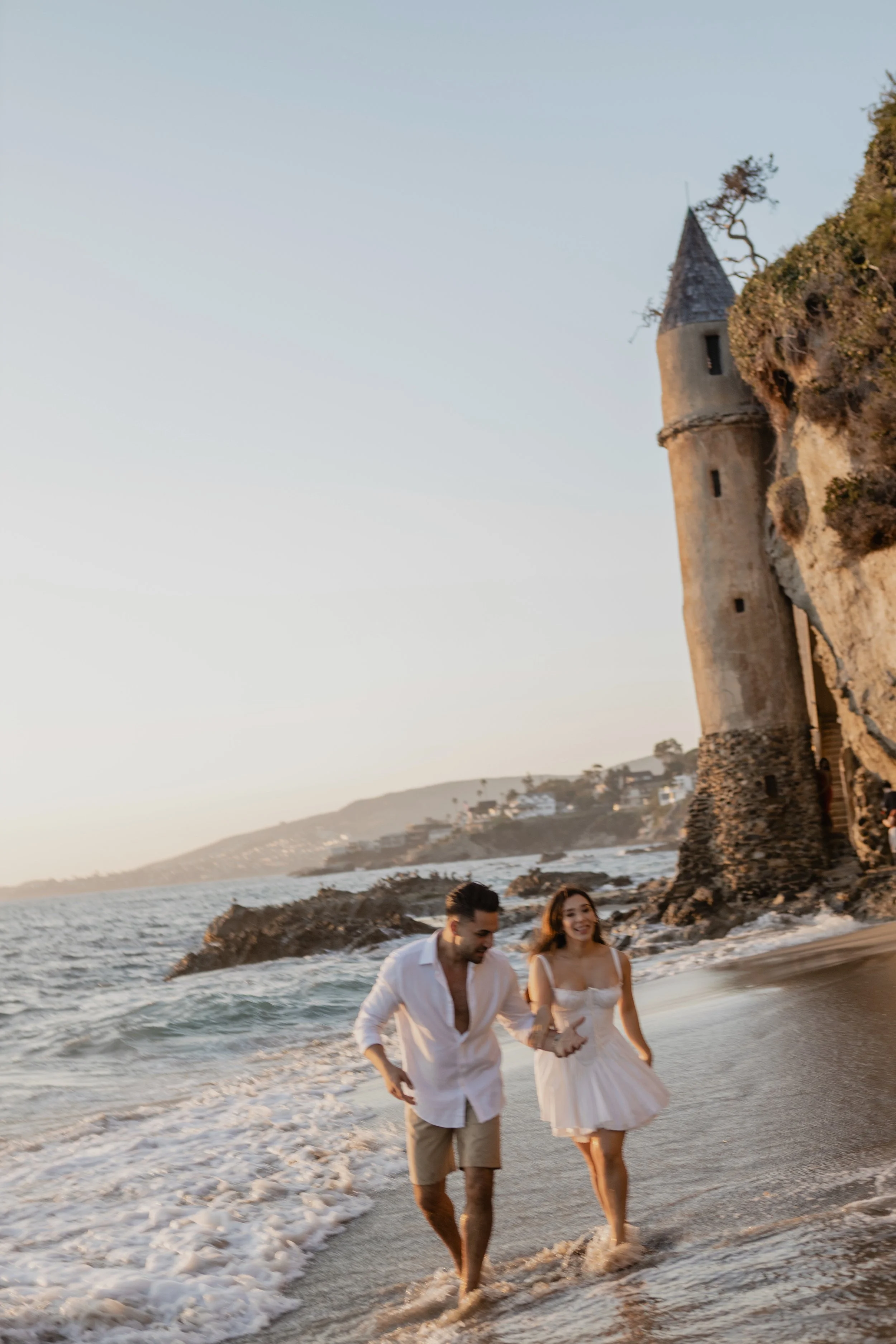 A couple walking along the shoreline of a beach with a large castle-like structure on a rocky cliff in the background. The man is dressed in a white shirt and khaki shorts, and the woman is wearing a white dress. The scene appears to be during sunset.