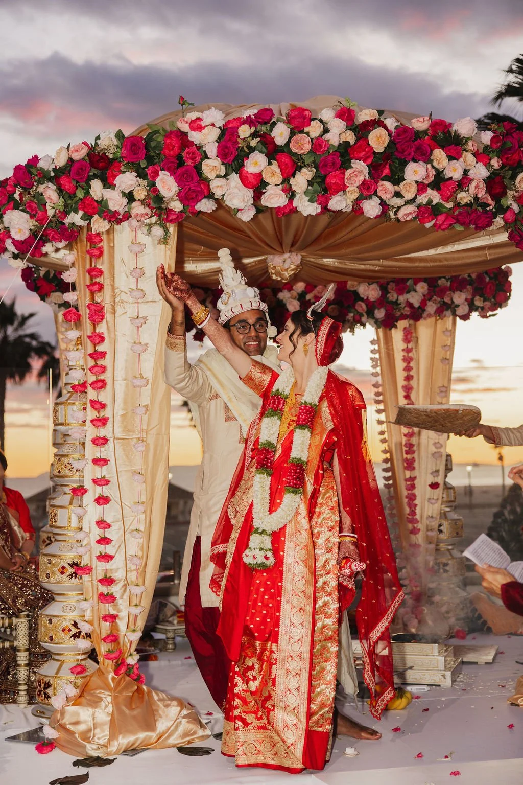 Sarah and Sanjit posing together beneath a red and pink floral mandap at The Waterfront Beach Resort in Huntington Beach