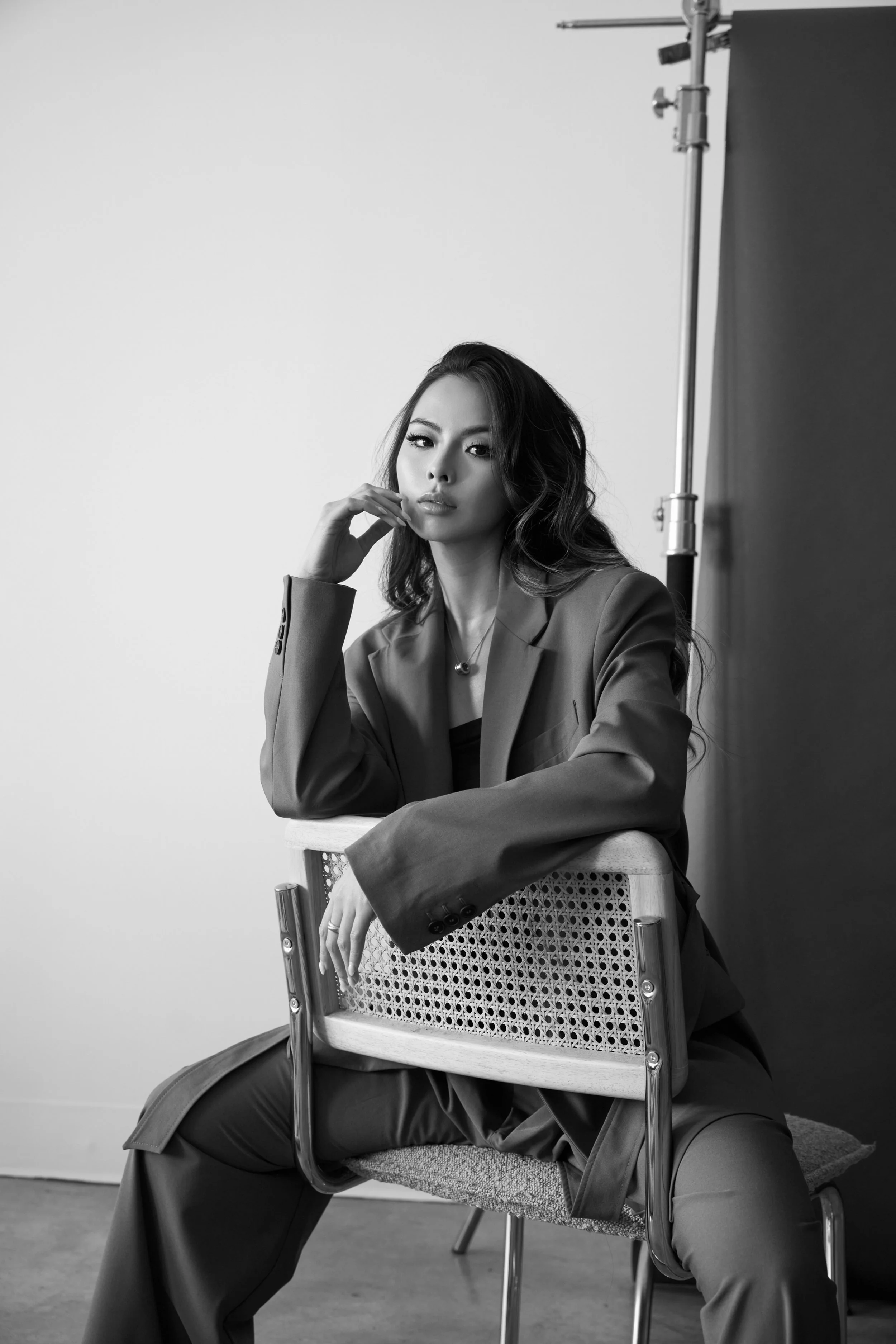 A woman sitting on a chair in a photography studio, looking at the camera. She has wavy hair and is wearing a suit. A backdrop stand and a dark backdrop are visible in the background, and she is posing with a relaxed expression.