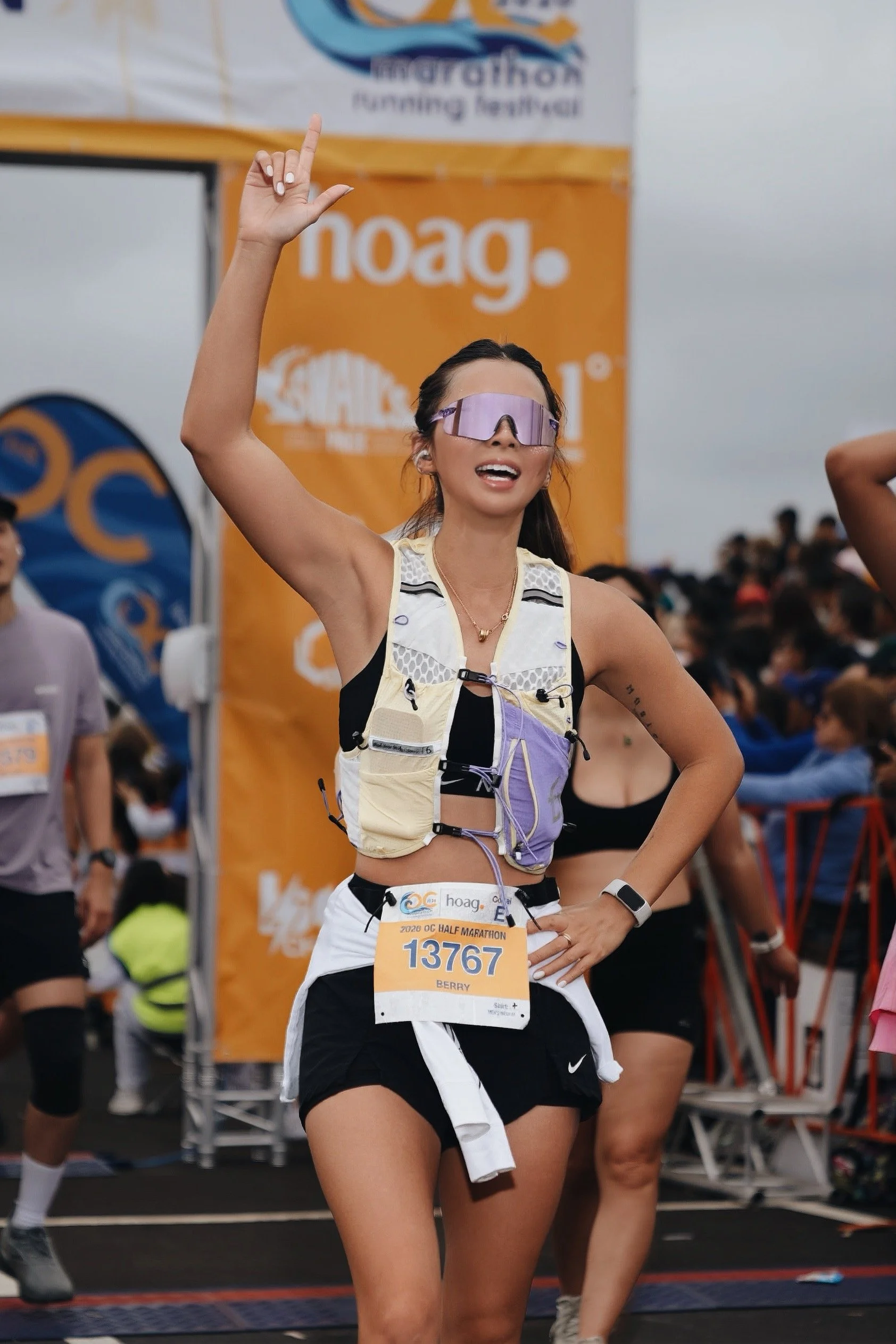 A woman running in a half marathon, wearing a bib number 13767, with her right hand raised and index finger pointing up. She is wearing sunglasses, a sports vest, and shorts. There are spectators and banners in the background.